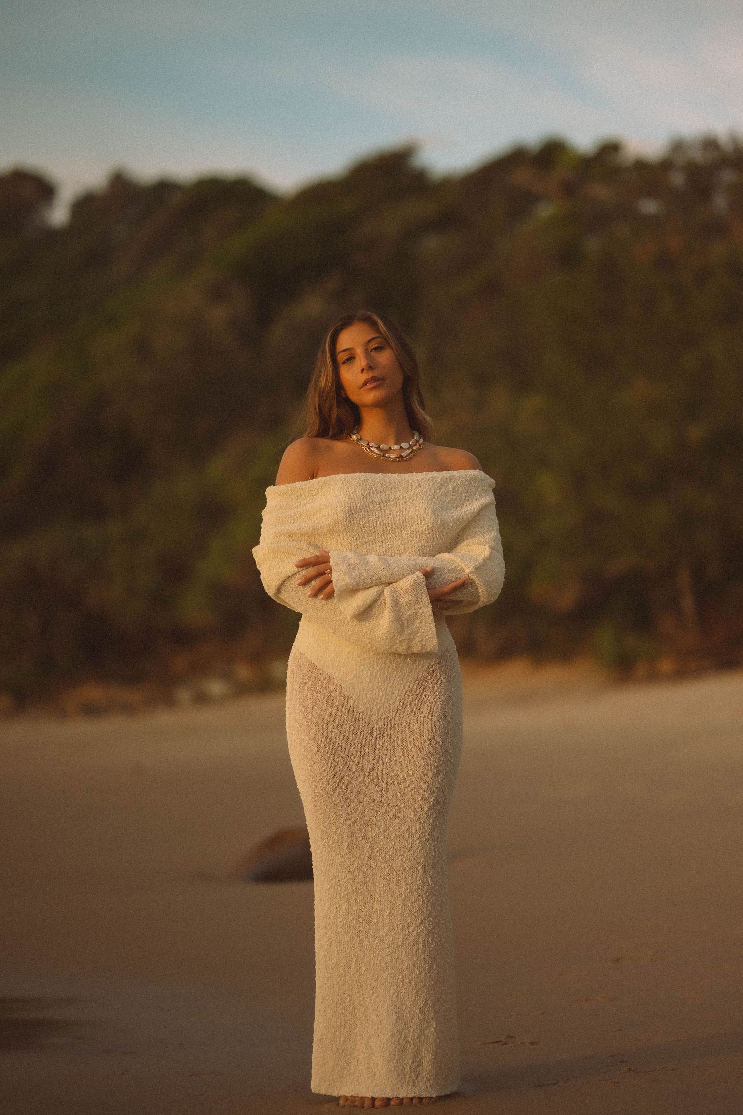 Woman in white off-shoulder knit dress and pearl necklace posing on a sandy beach at golden hour.
