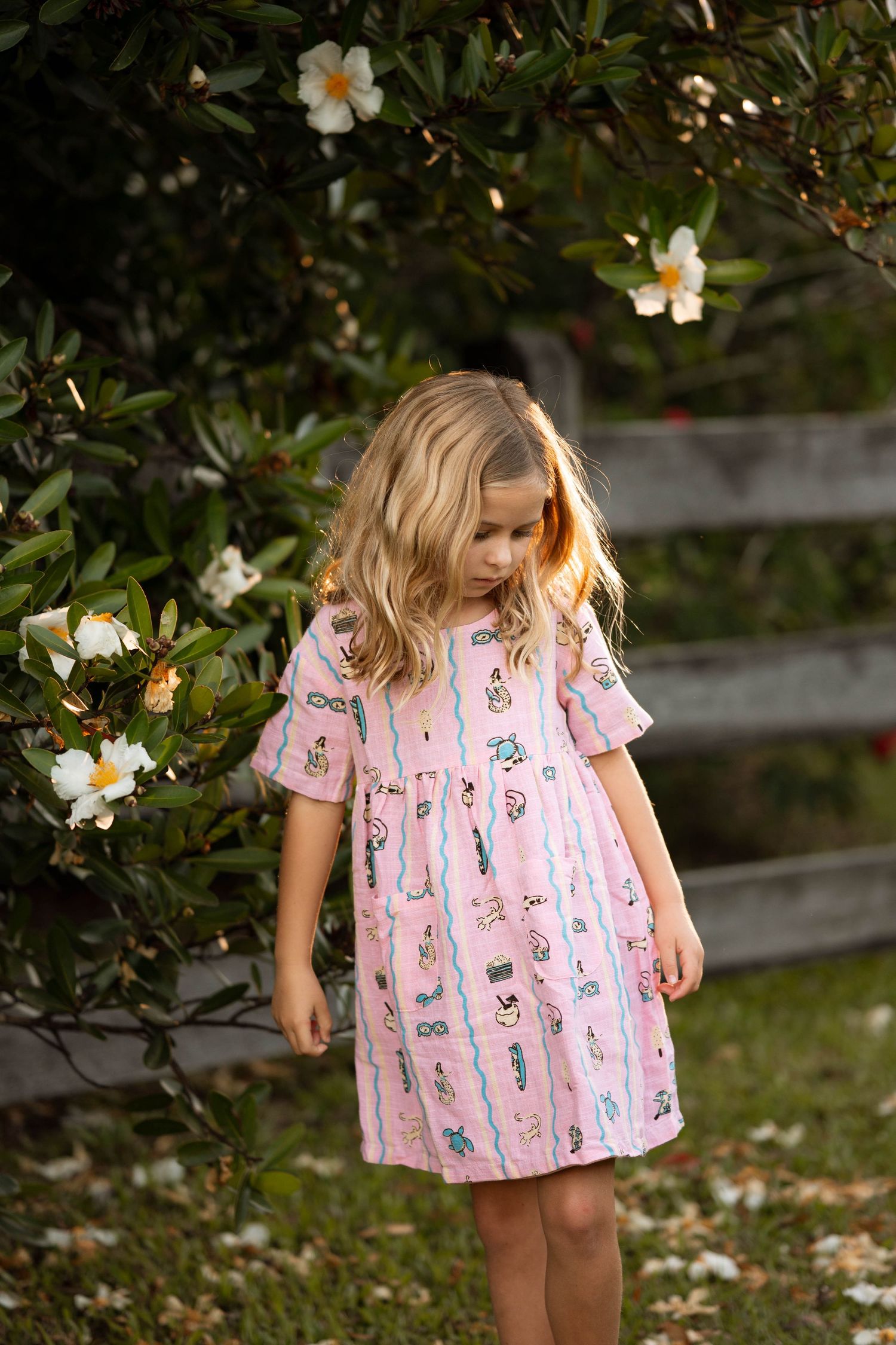 Young girl in pink printed dress standing near blooming white flowers in a garden setting.