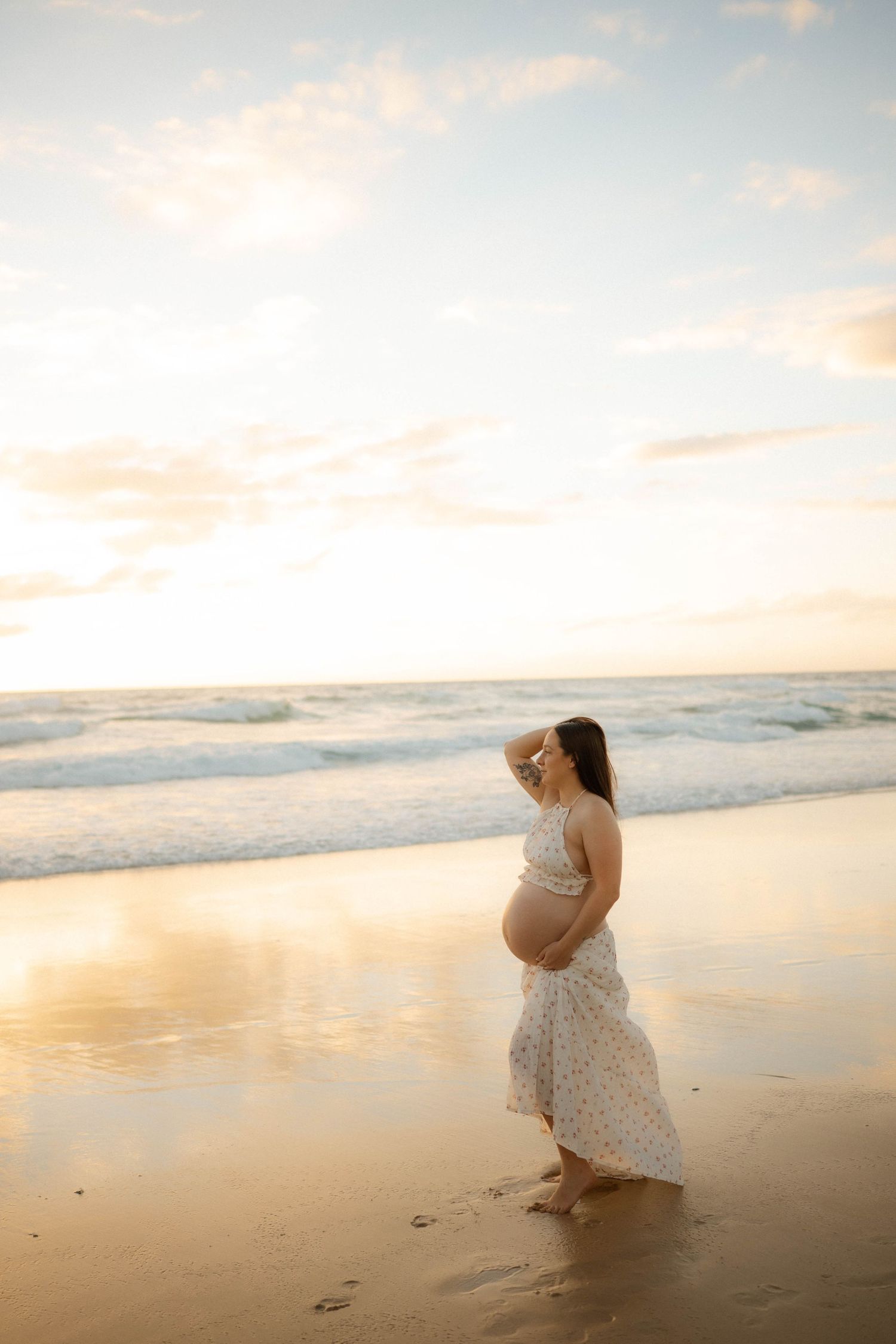 Pregnant woman in floral dress standing on beach at golden sunset, waves reflecting warm light on wet sand.