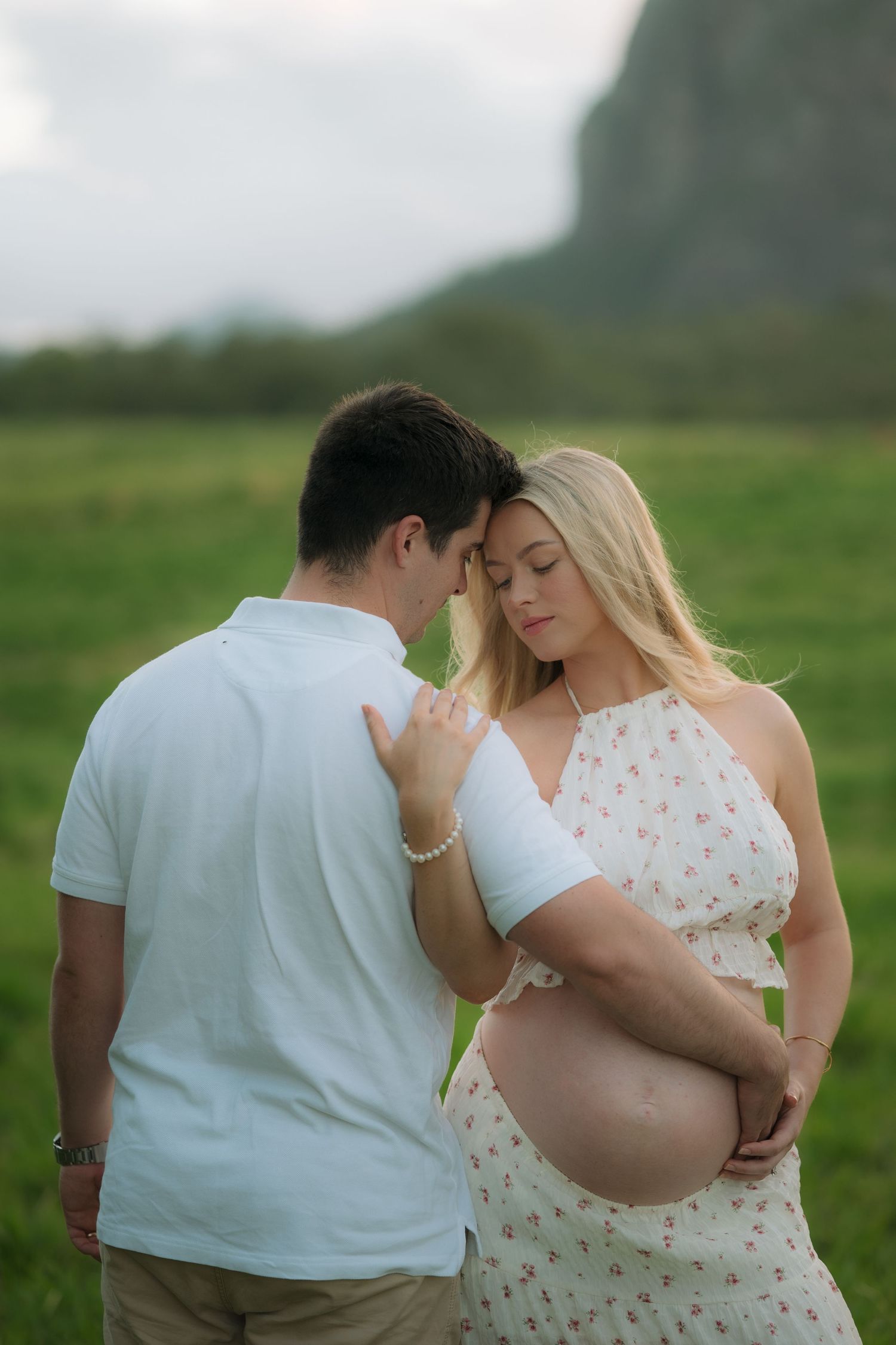 Tender moment as couple leans foreheads together in green field, woman cradling baby bump with mountain behind.
