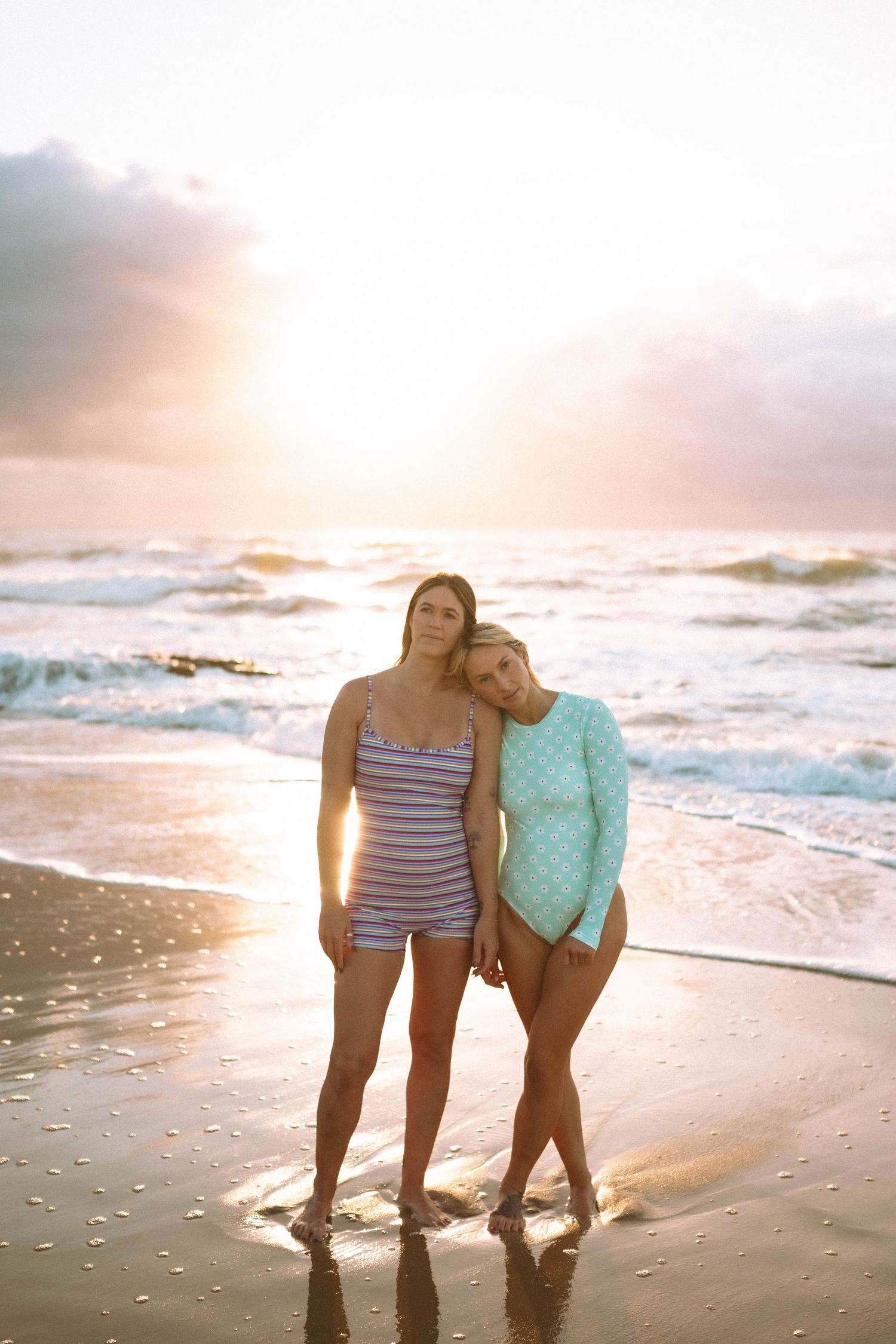 Two women in swimsuits standing together on beach shore bathed in warm golden sunset light.