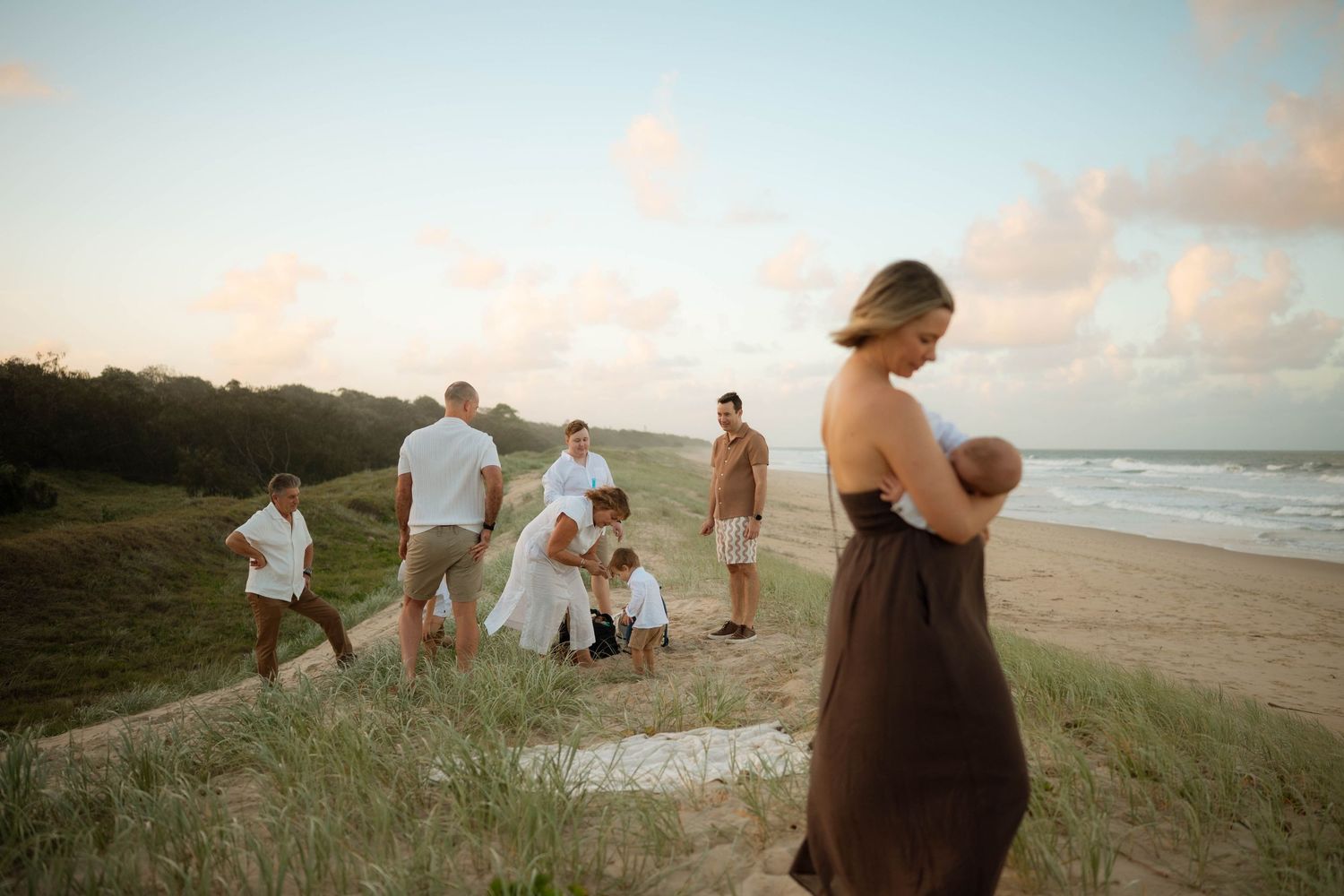 Extended family gathering on coastal sand dunes at sunset with woman holding baby.