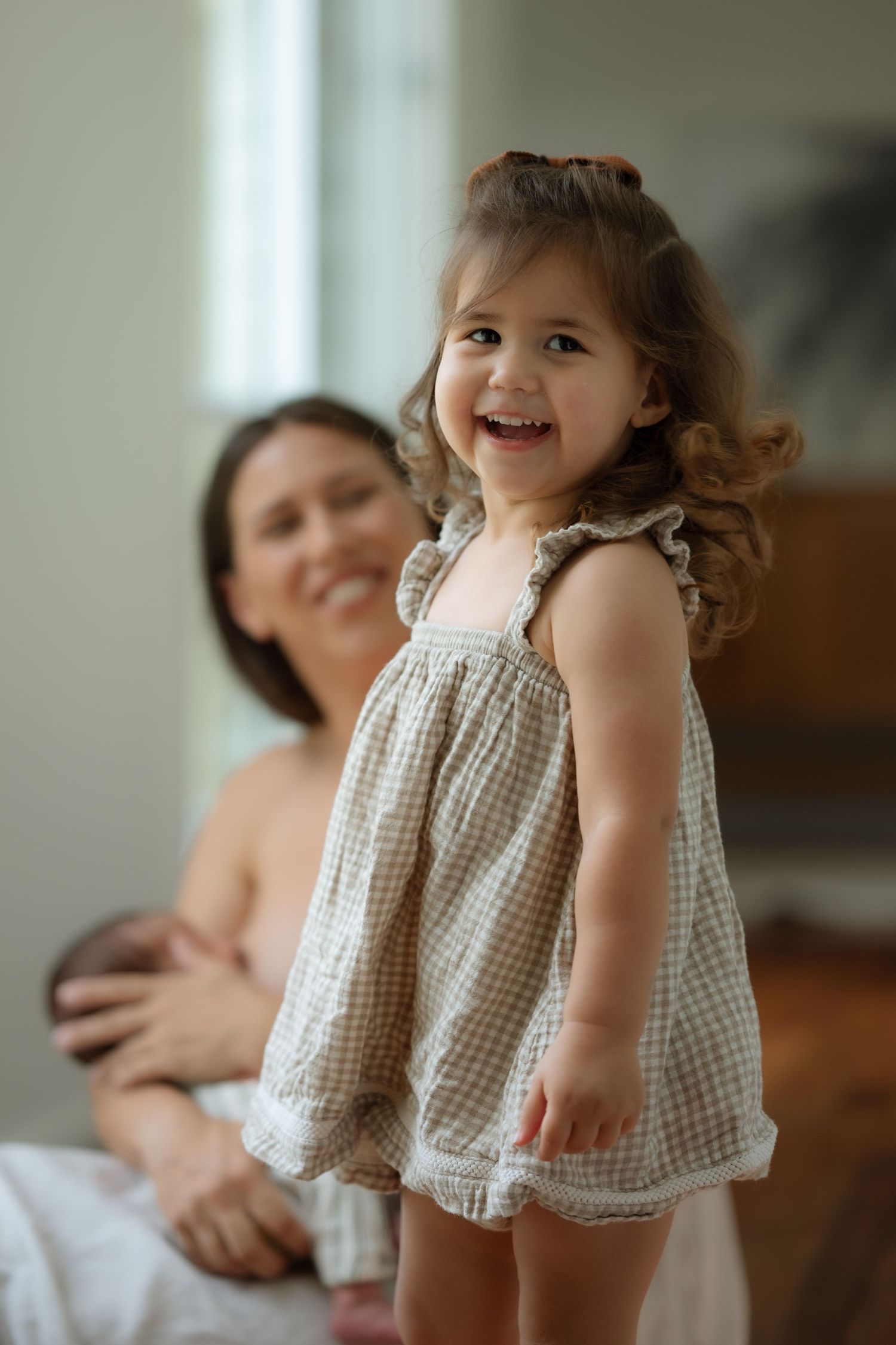 Smiling curly-haired toddler girl in gingham dress standing in front of blurred mother with newborn.