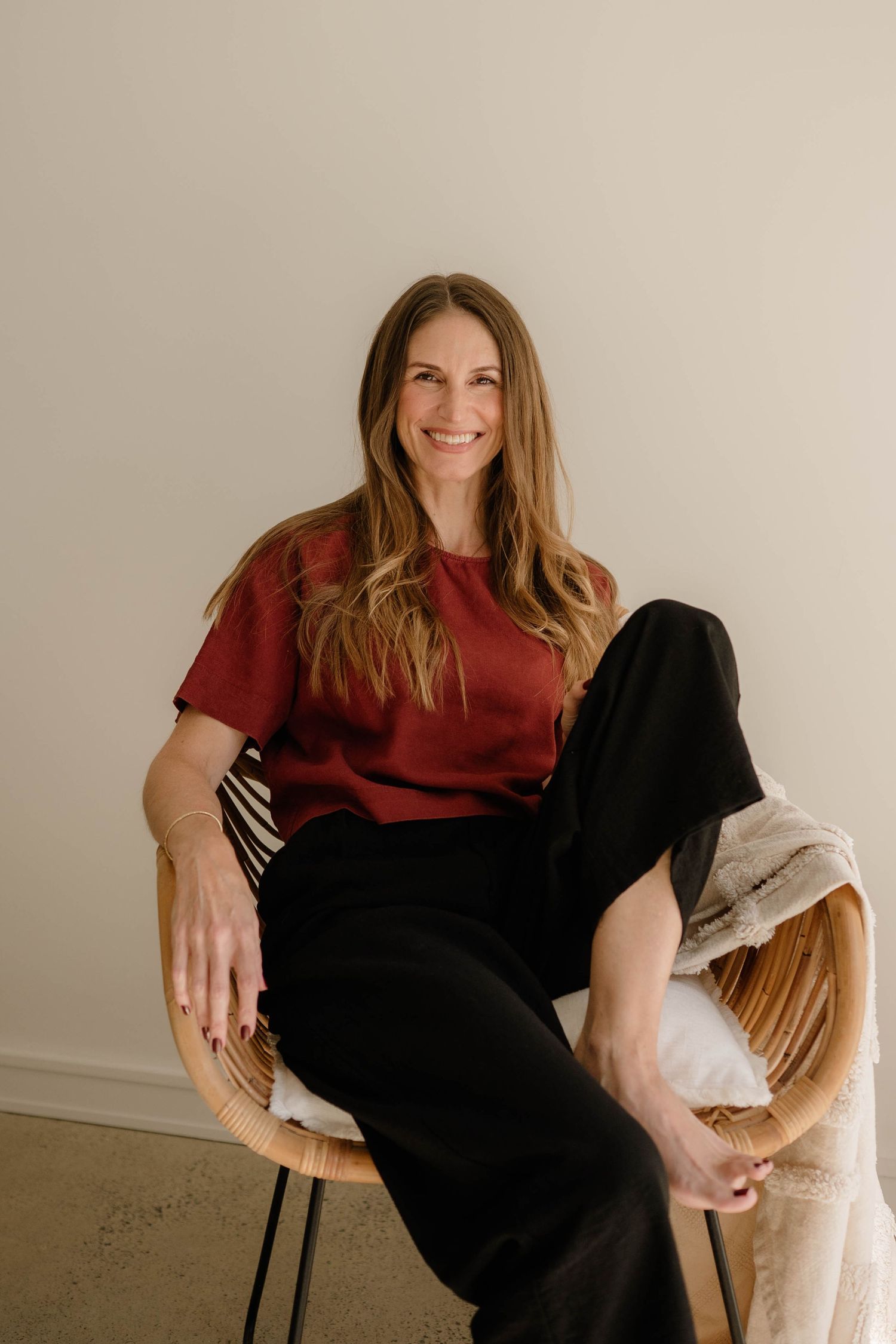 Smiling woman in red top and black pants relaxing in modern rattan chair indoors.