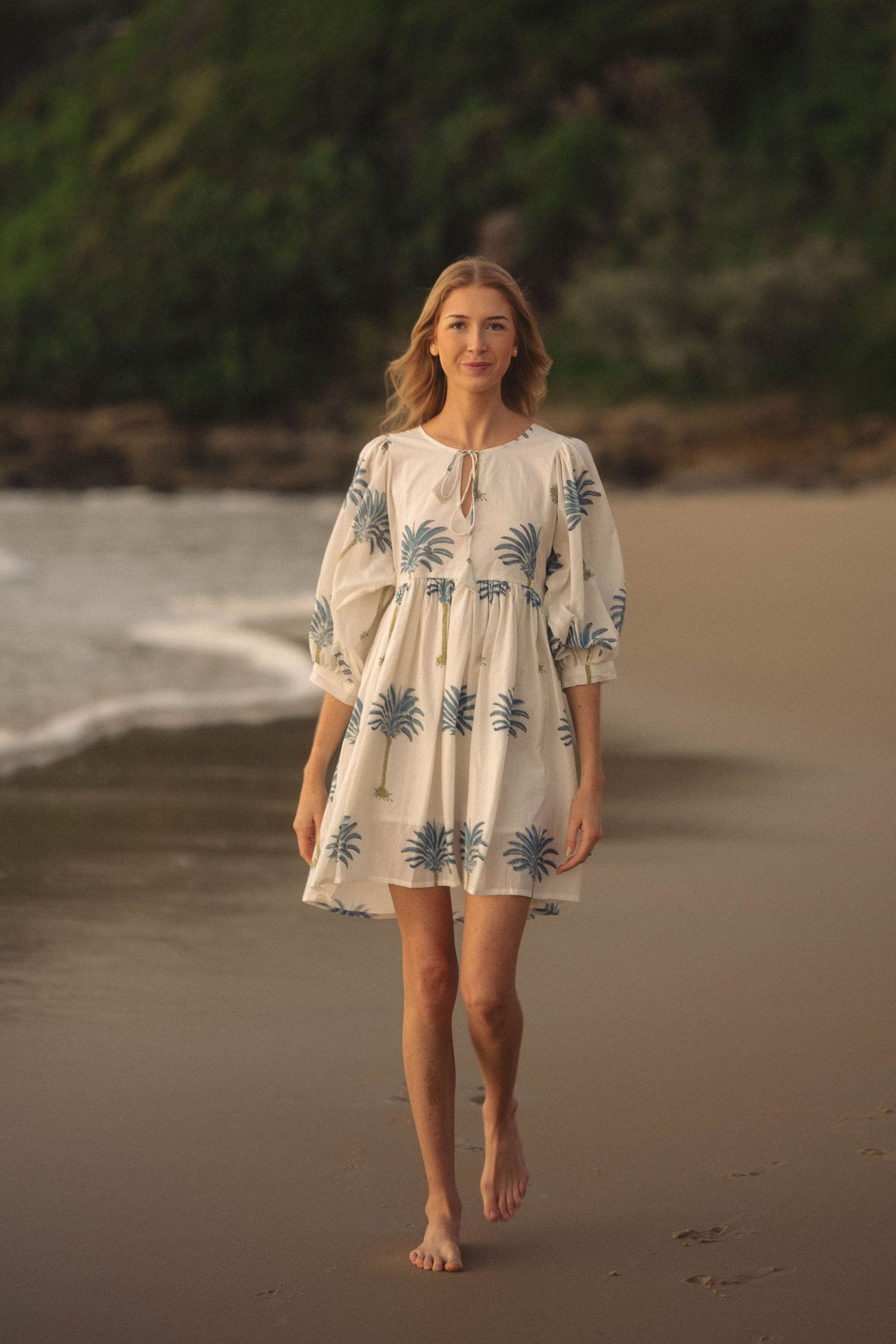 Woman in white blue palm print dress walking barefoot on sandy beach with ocean waves.