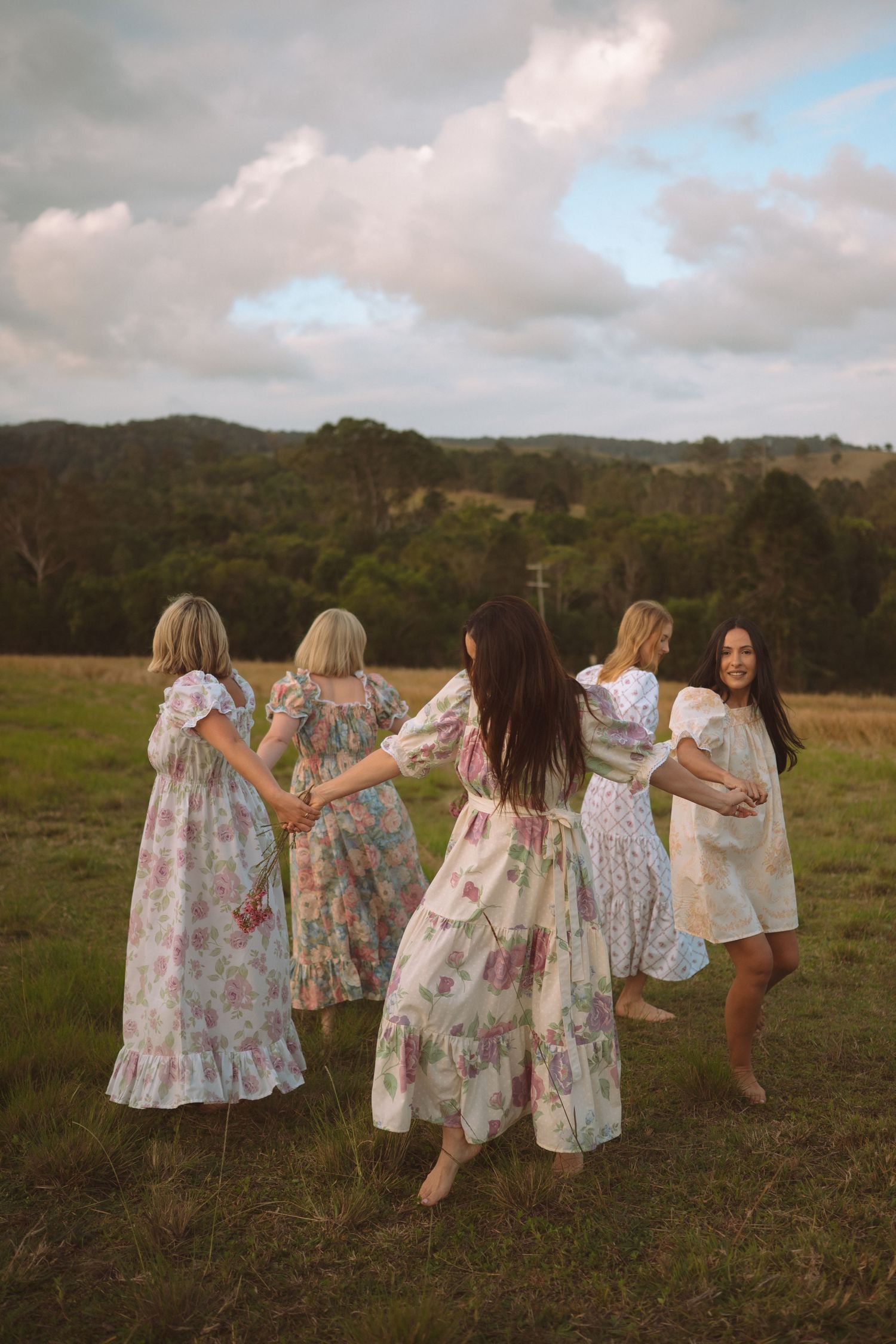 Five women in floral dresses hold hands in a circle on a green field with rolling hills.