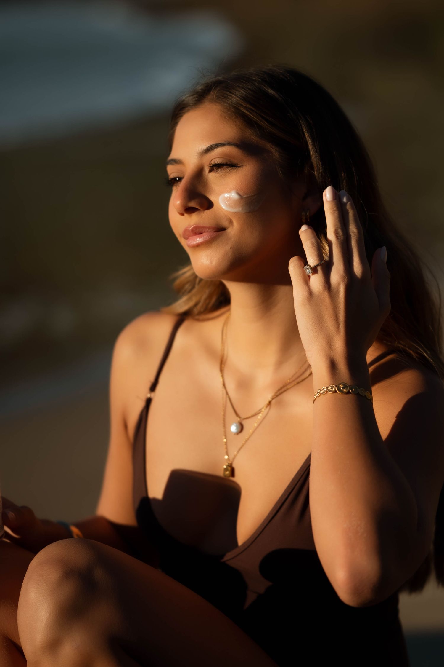 Woman applying sunscreen to cheek on beach at golden hour wearing jewelry and brown swimsuit.