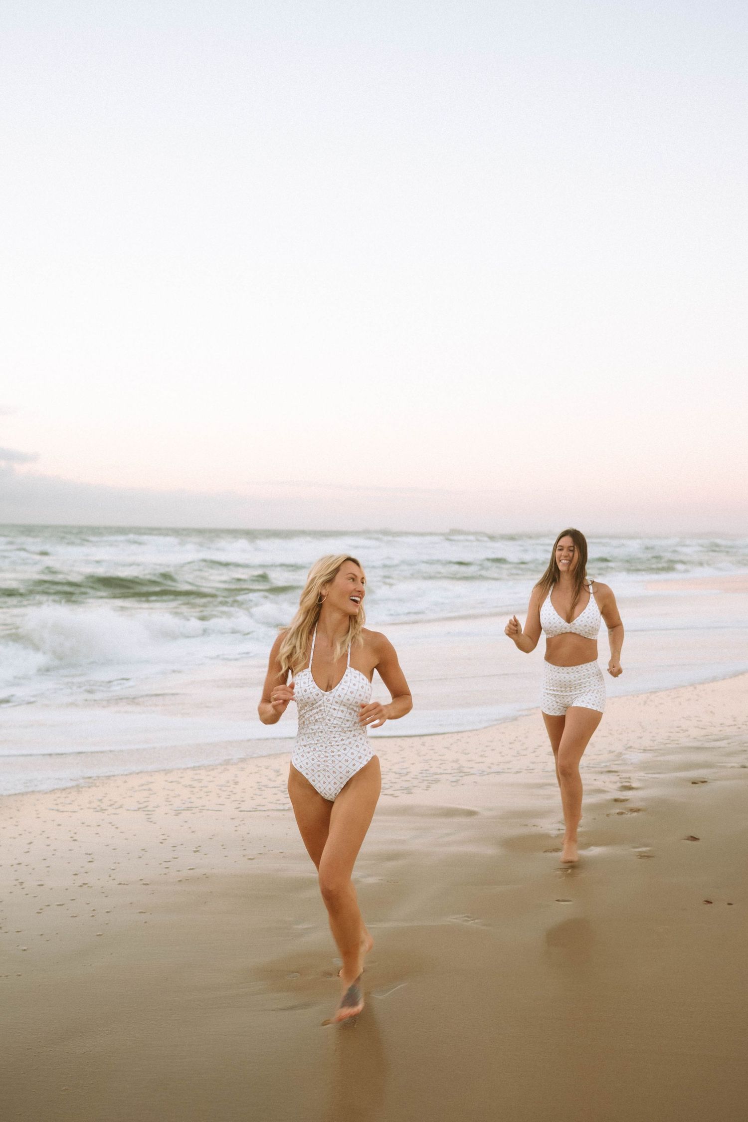 Two women in white swimsuits running joyfully along a sandy beach at sunset with ocean waves behind them.