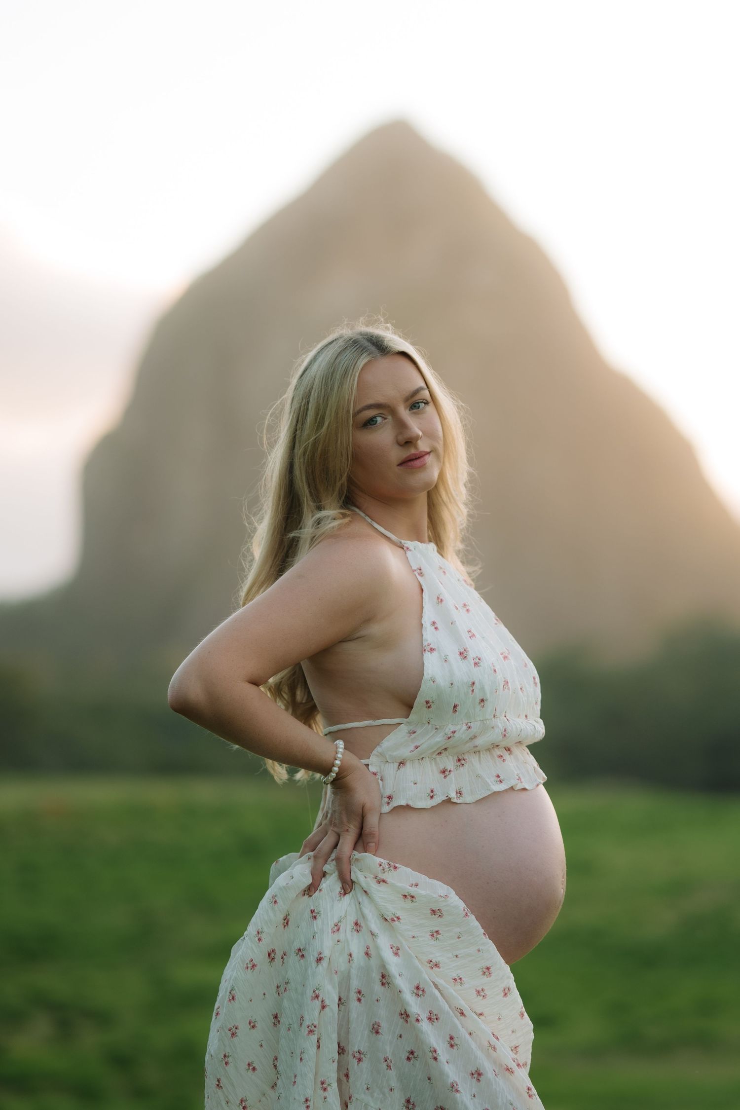 Blonde pregnant woman in floral maternity dress poses with hand on hip before a scenic rock mountain at golden hour.