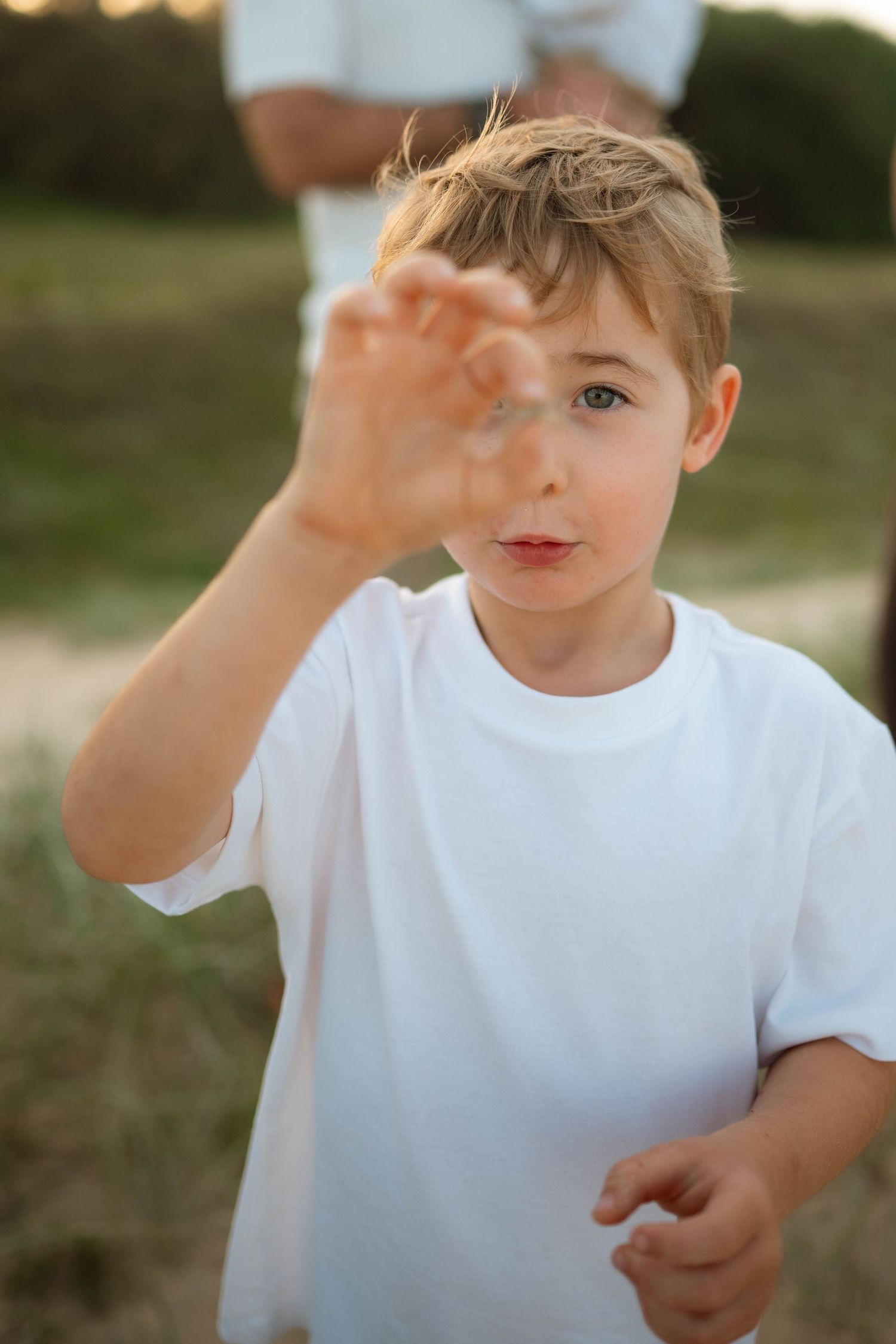 Blond boy in white t-shirt reaching toward camera outdoors in golden light.