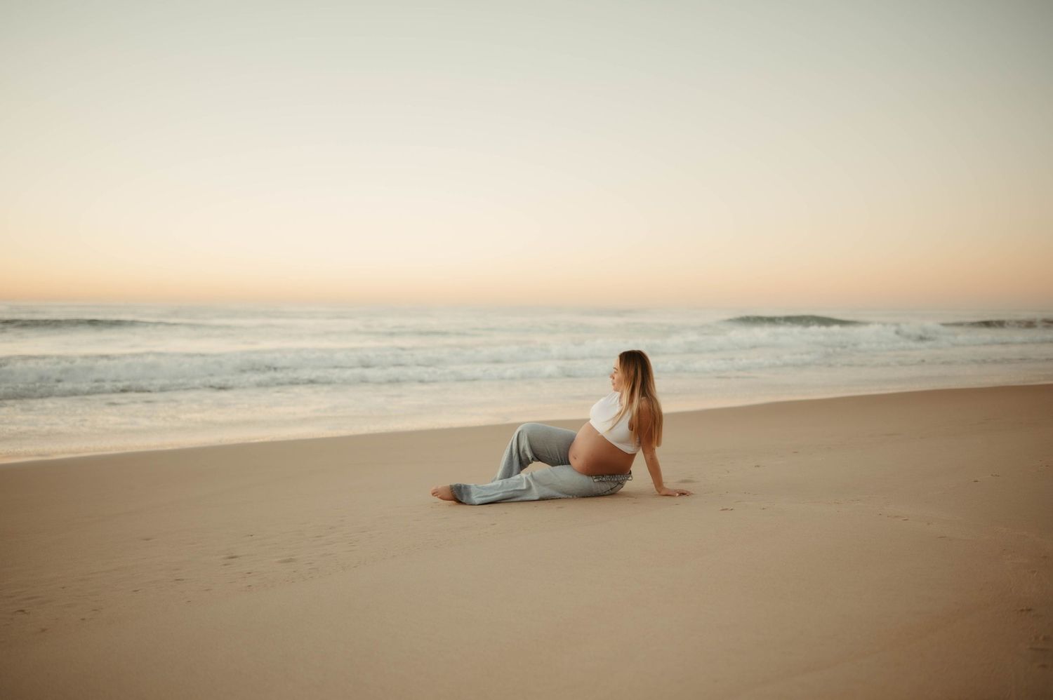Pregnant woman in jeans sitting gracefully on a serene beach at pastel sunrise.
