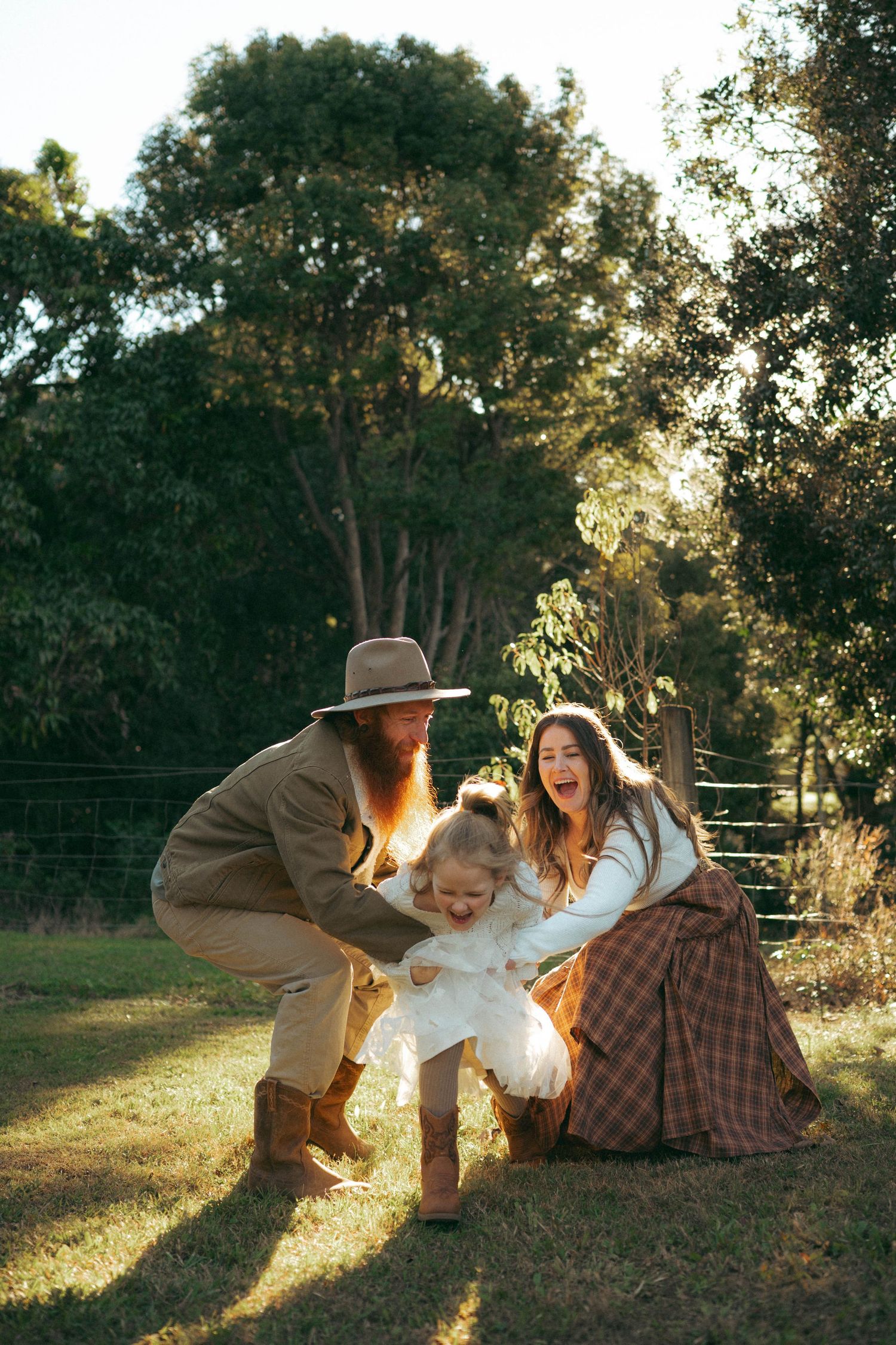 Joyful family playing with young child on grass with sunlit trees in background.