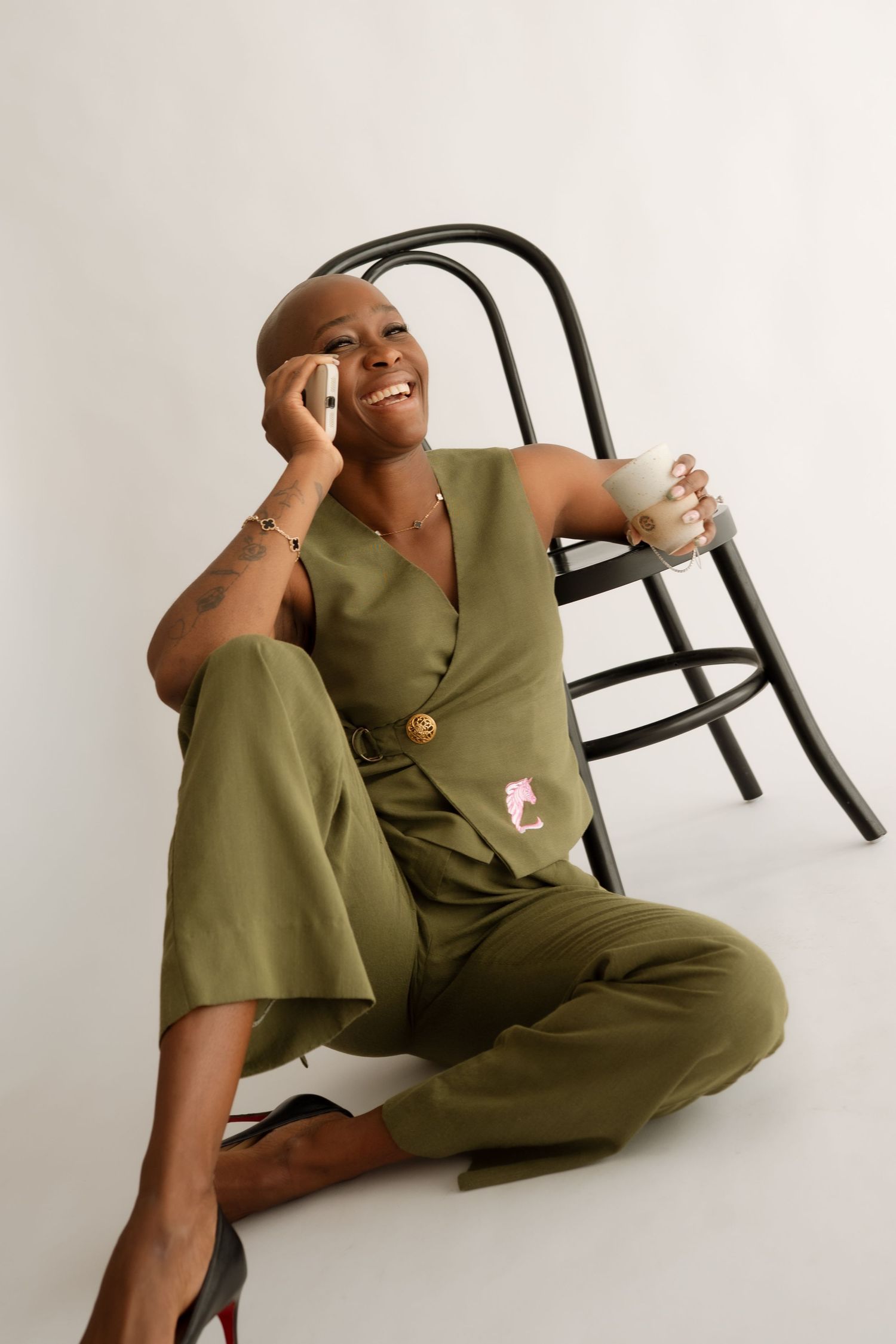 Smiling bald woman in olive green jumpsuit sitting on floor, talking on phone while holding a coffee cup.