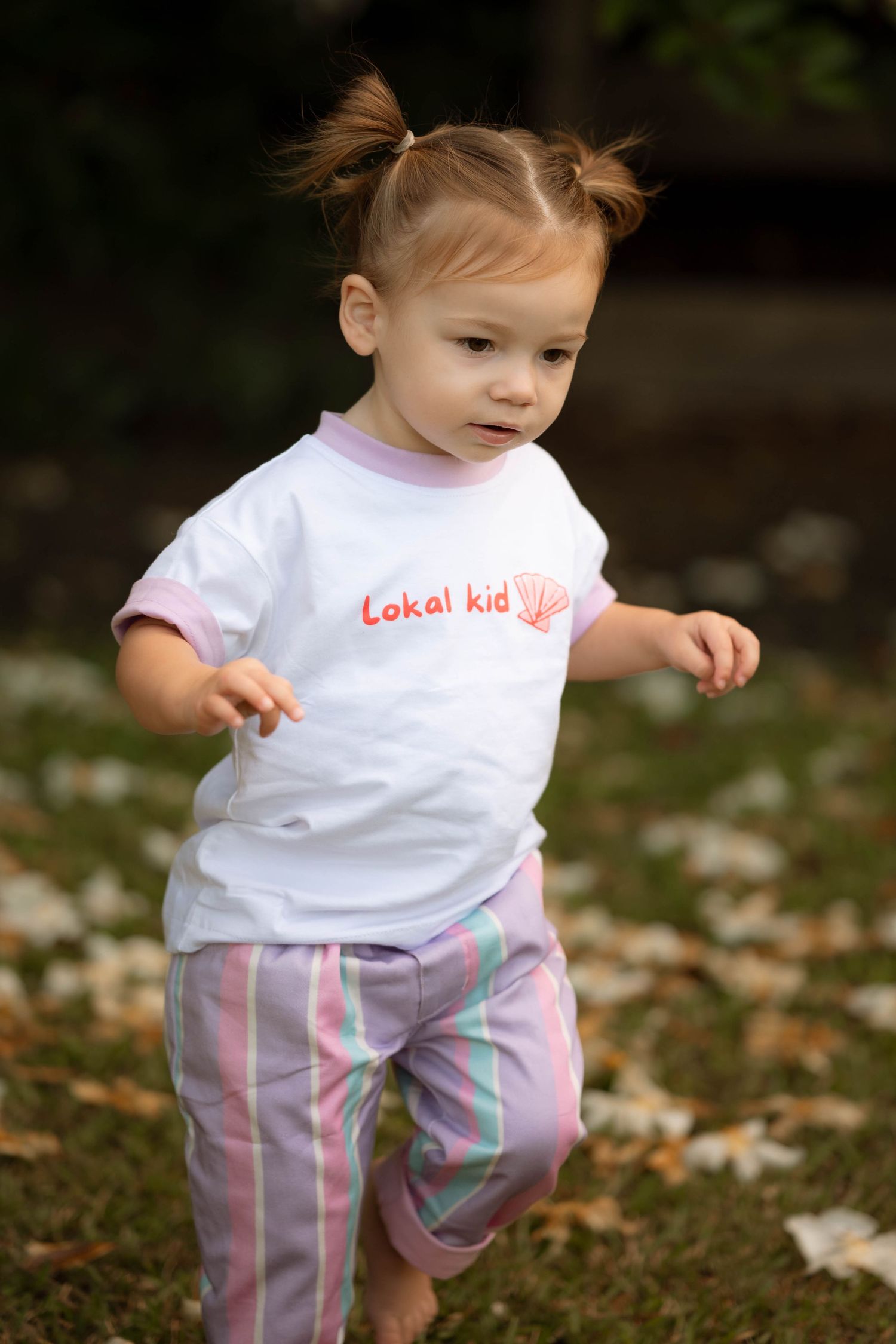 Toddler girl in 'Lokal Kid' white tee and striped pants playing outdoors on grass.