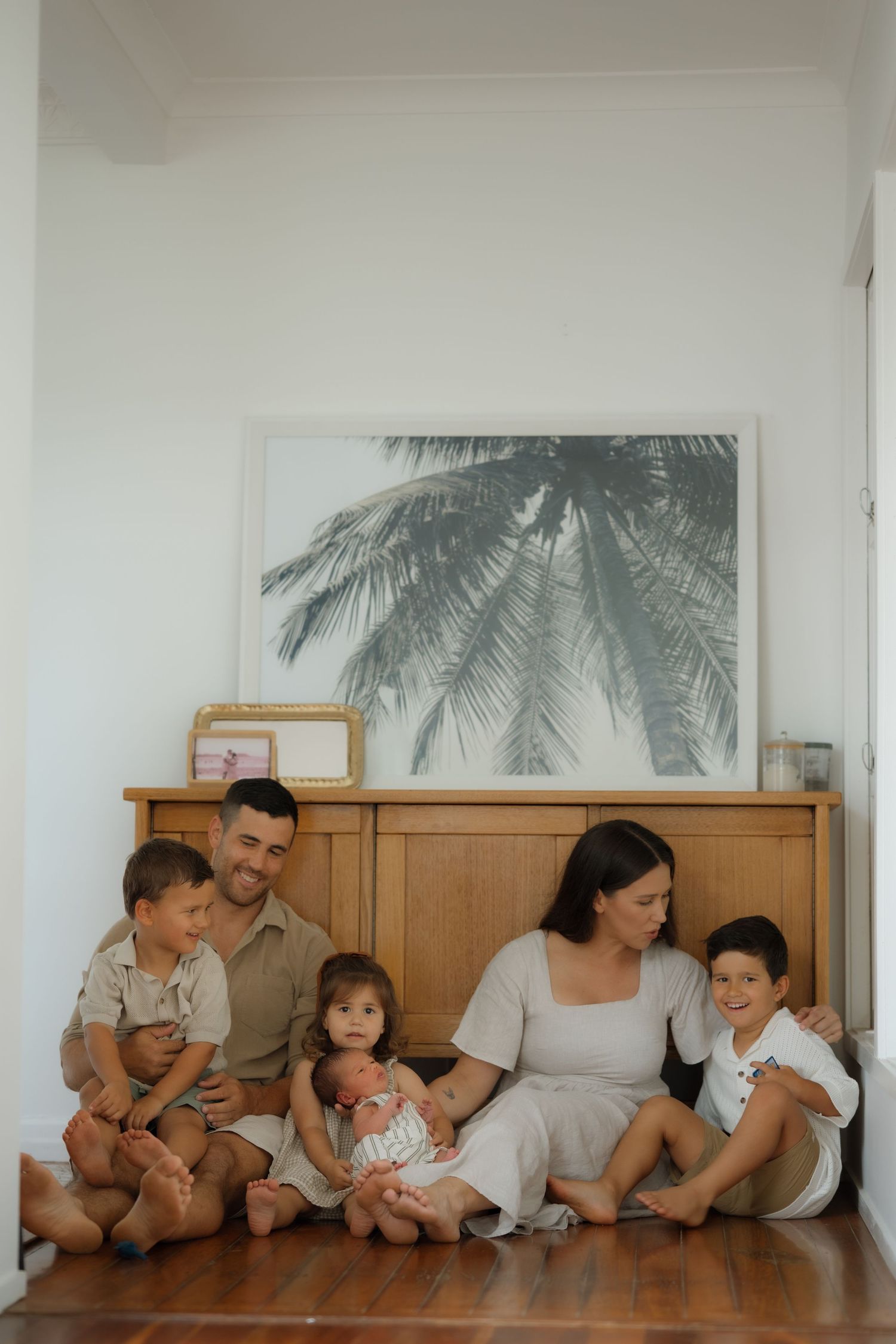 Family of six sitting on floor against wooden dresser with palm tree artwork, parents with three kids and newborn baby.