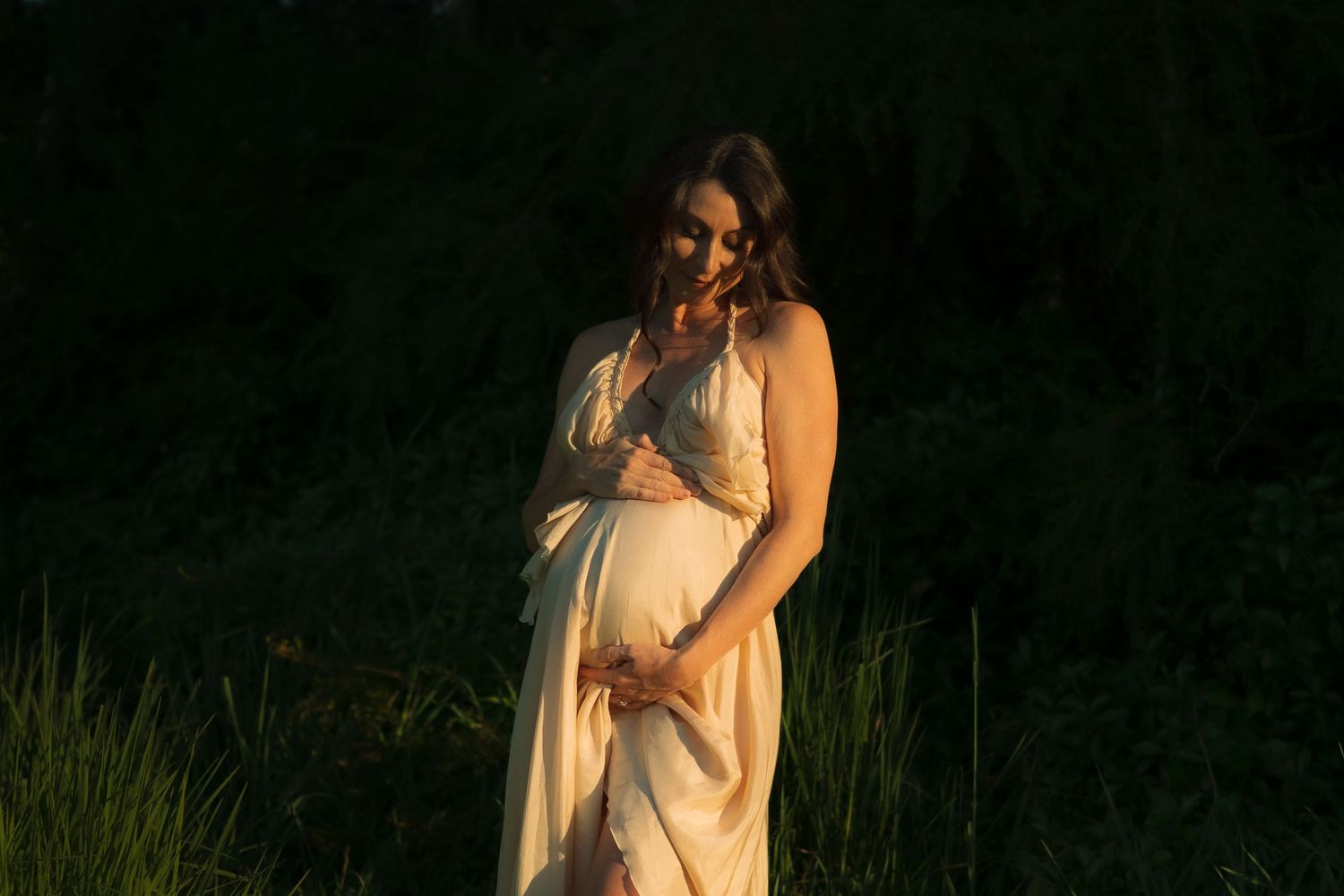 Expectant mother in flowing cream gown tenderly cradles baby bump in warm sunset light outdoors.