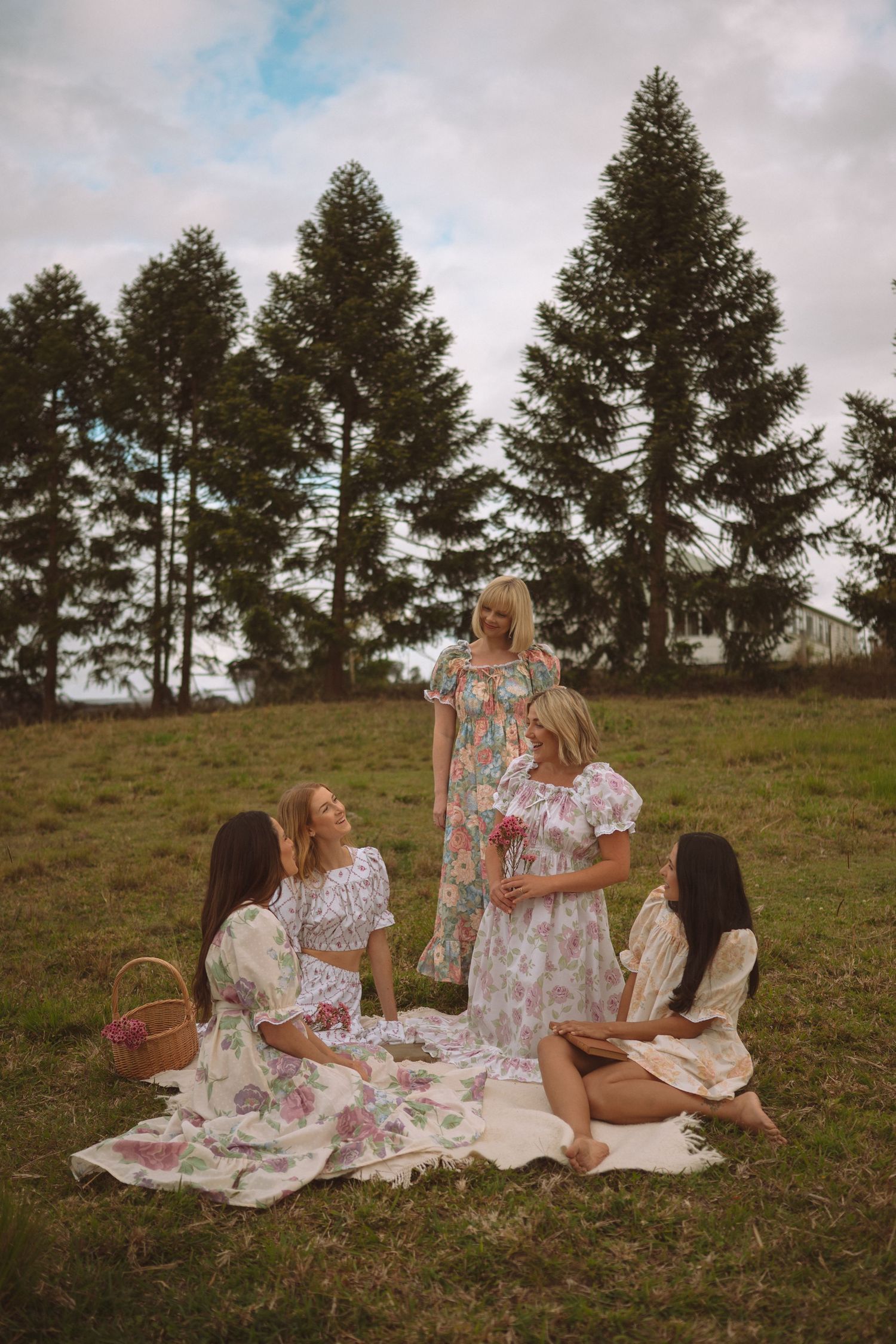 Four women in floral dresses enjoy a picnic on a blanket surrounded by tall pine trees on a green hillside.