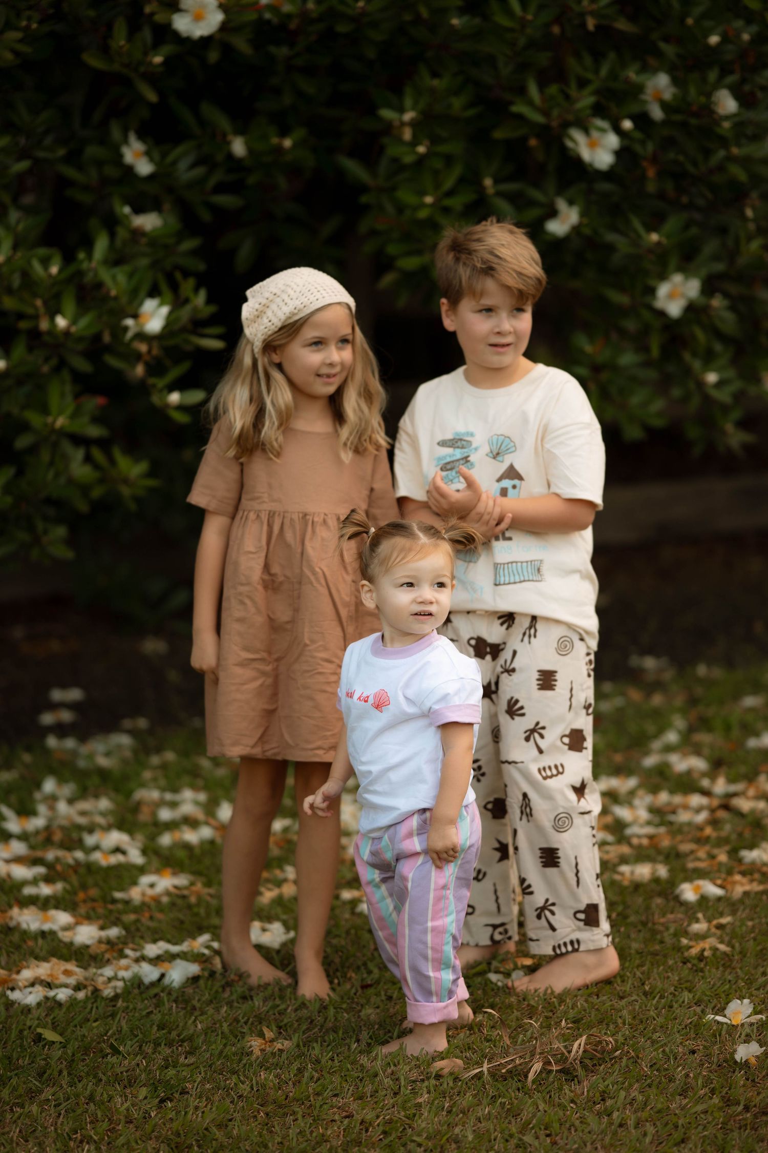 Three barefoot children pose outdoors on grass near flowering bushes, wearing casual summer outfits.