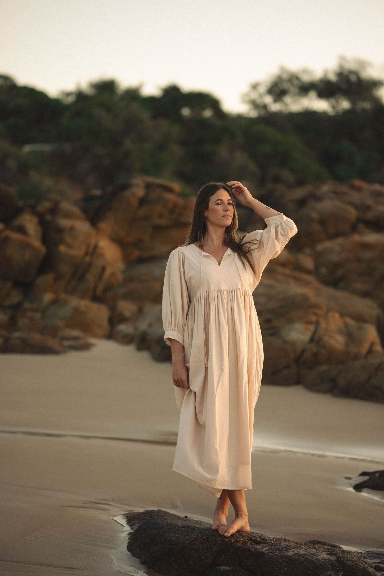 Woman in flowing cream dress stands barefoot on coastal rocks at sunset with trees in background.