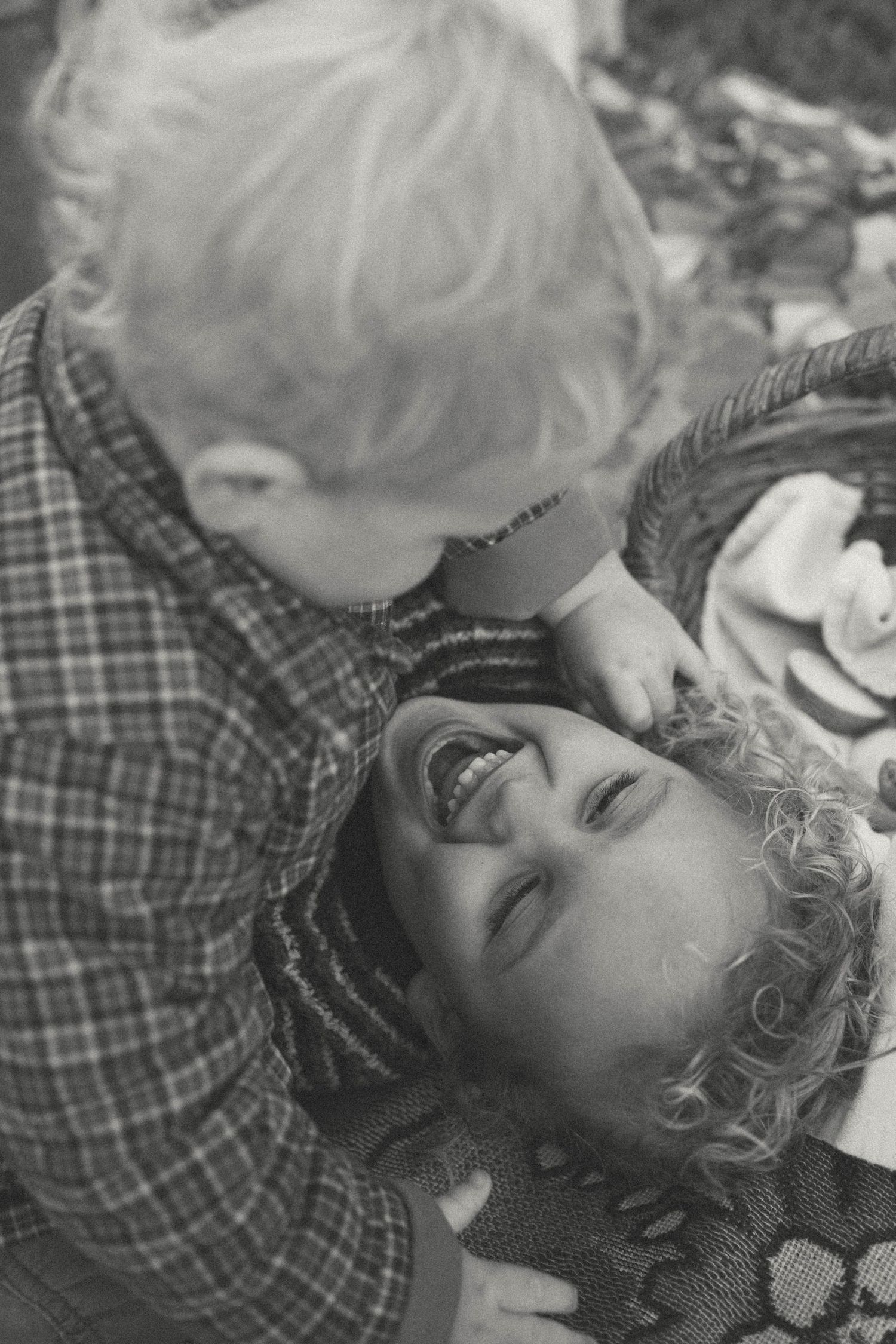 Black and white photo of a laughing curly-haired child being kissed by a young blonde toddler near a wicker basket.