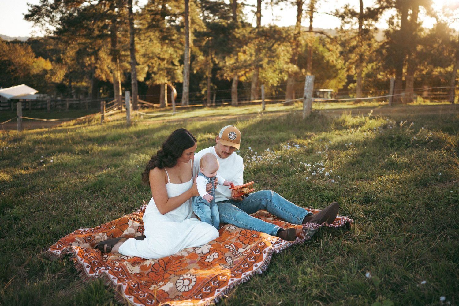 Family sitting on a blanket in a sunny field at golden hour, parents with baby enjoying an outdoor picnic.