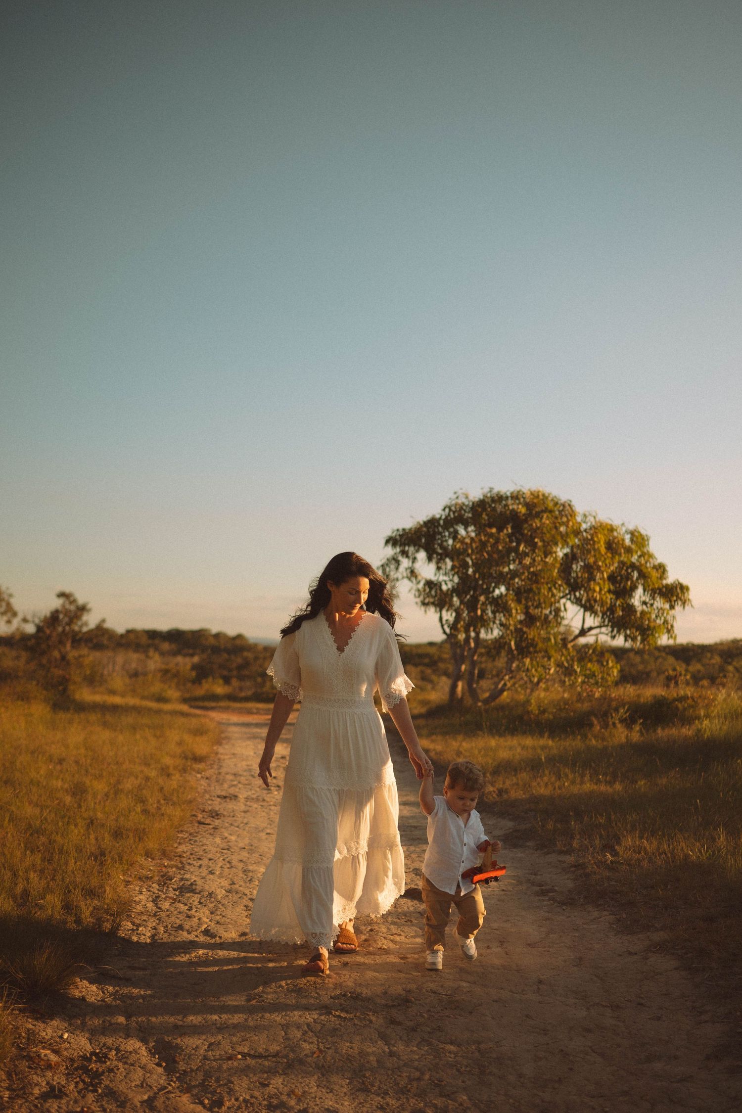 A woman in a white dress walks hand-in-hand with a toddler on a sunny dirt path surrounded by golden fields.