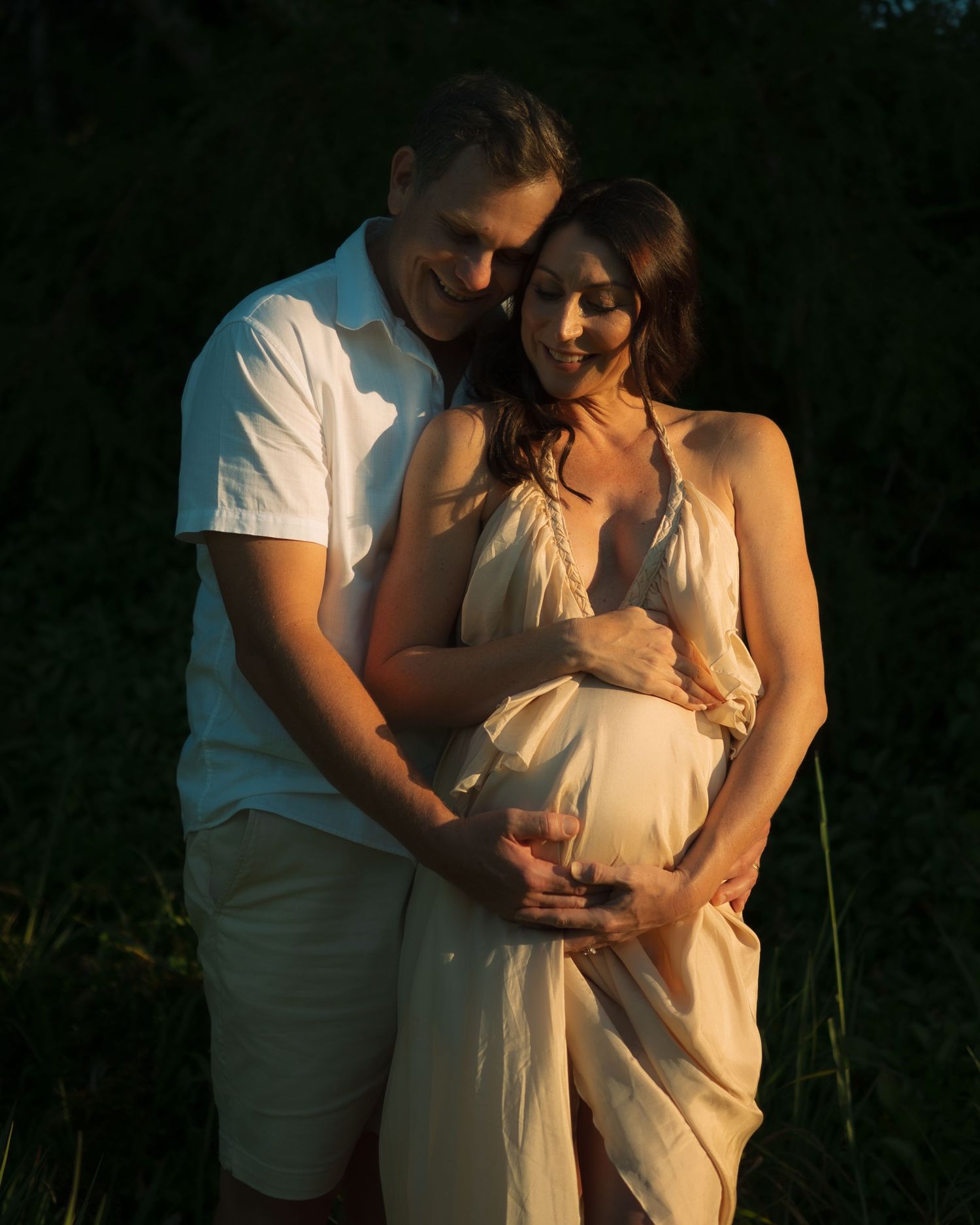 Loving couple embraces outdoors at sunset, hands cradling pregnant belly in warm golden light.