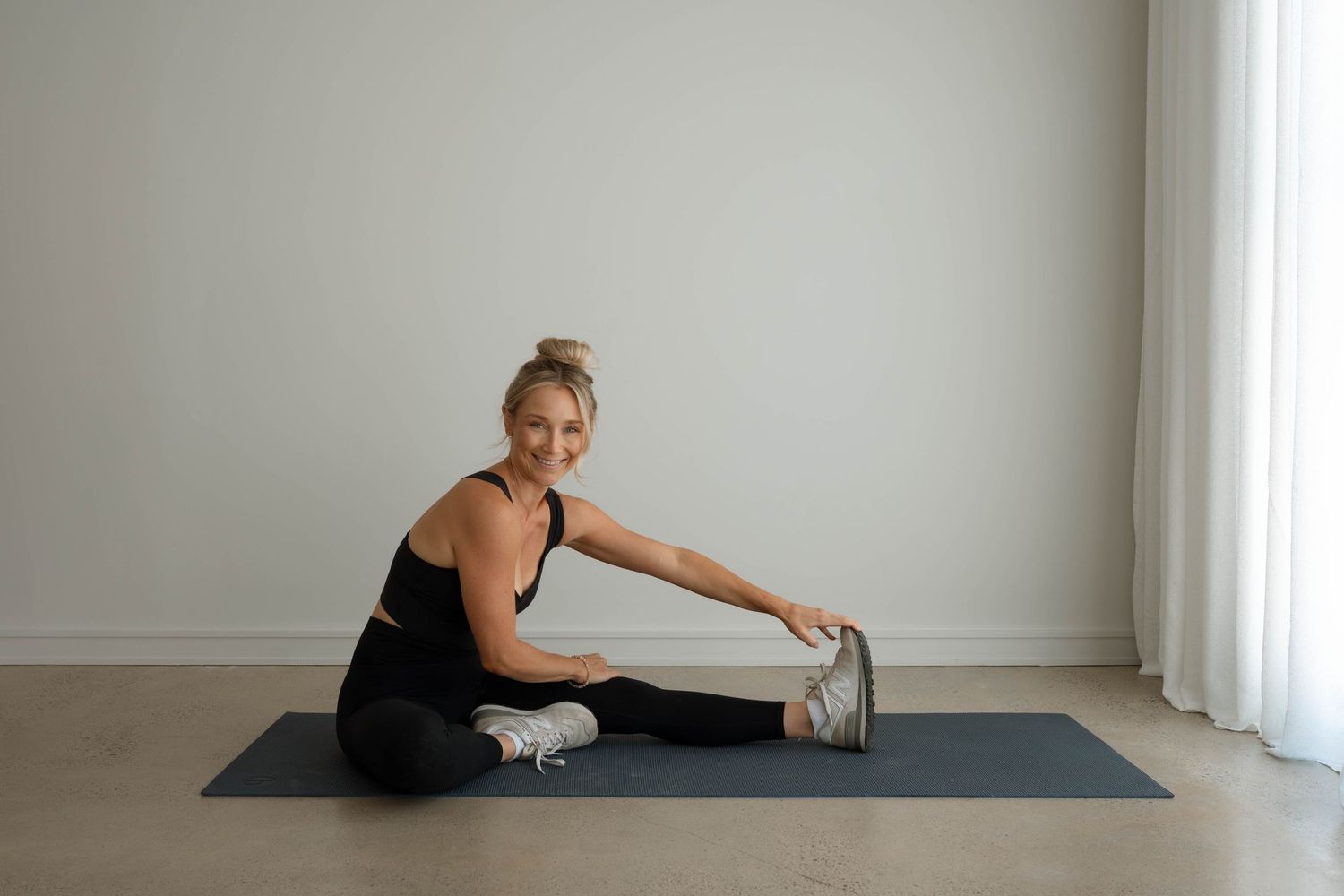 Smiling woman in black activewear stretching forward on yoga mat in bright minimal studio.