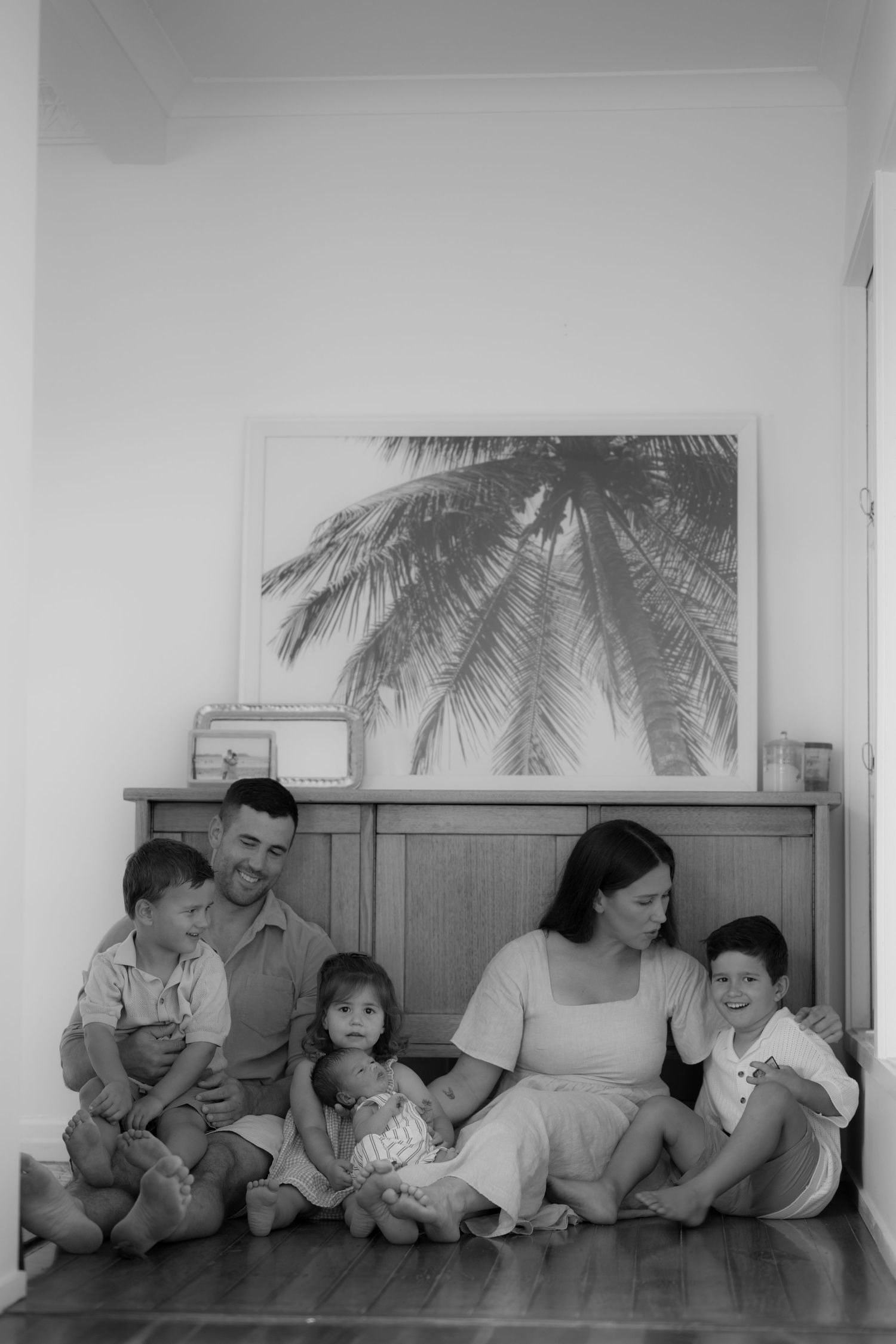 Black and white family portrait of parents with three children and newborn sitting on floor by wooden dresser.
