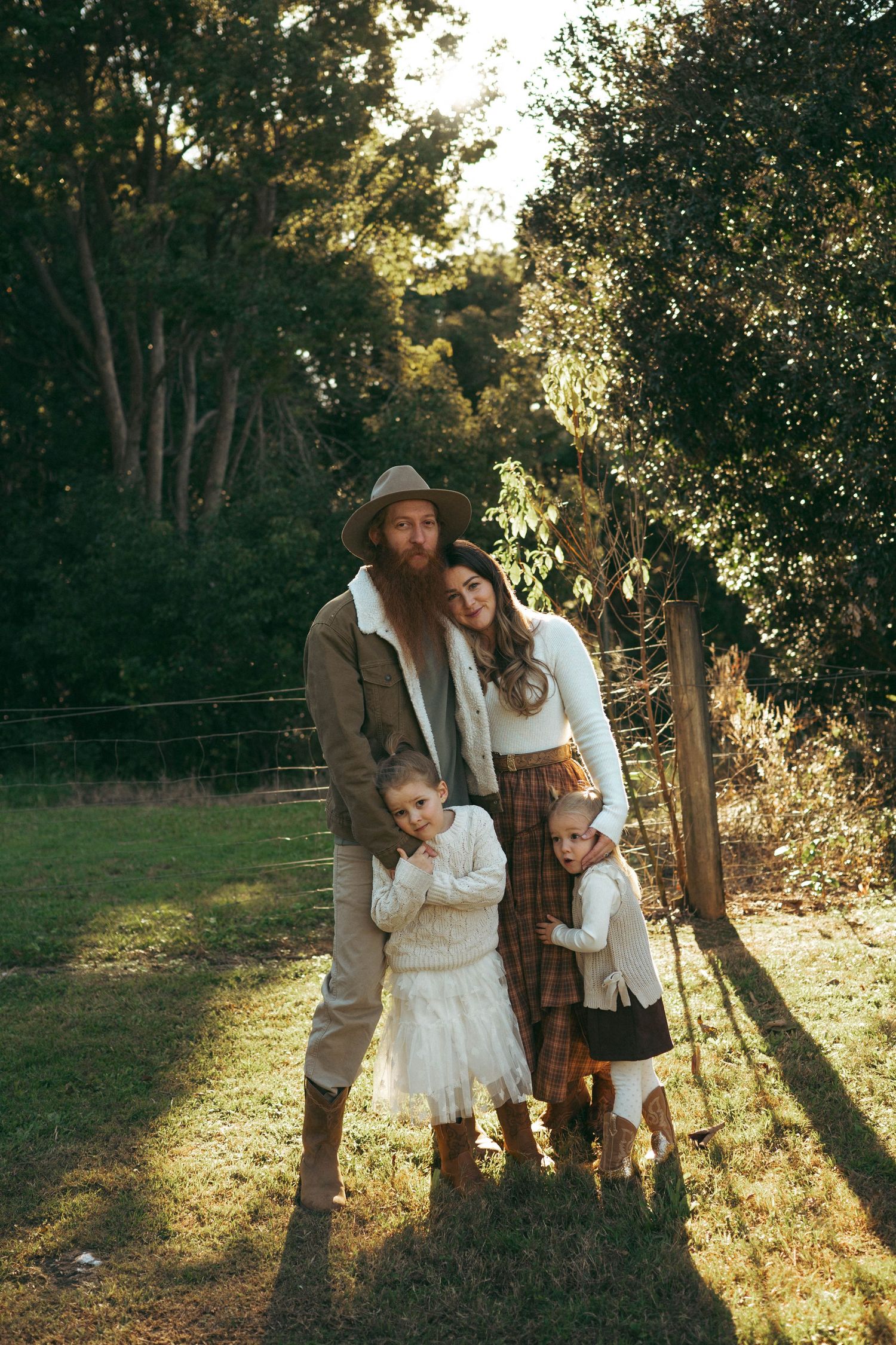 Boho family portrait outdoors in warm sunlight, parents and two children by a rustic fence.