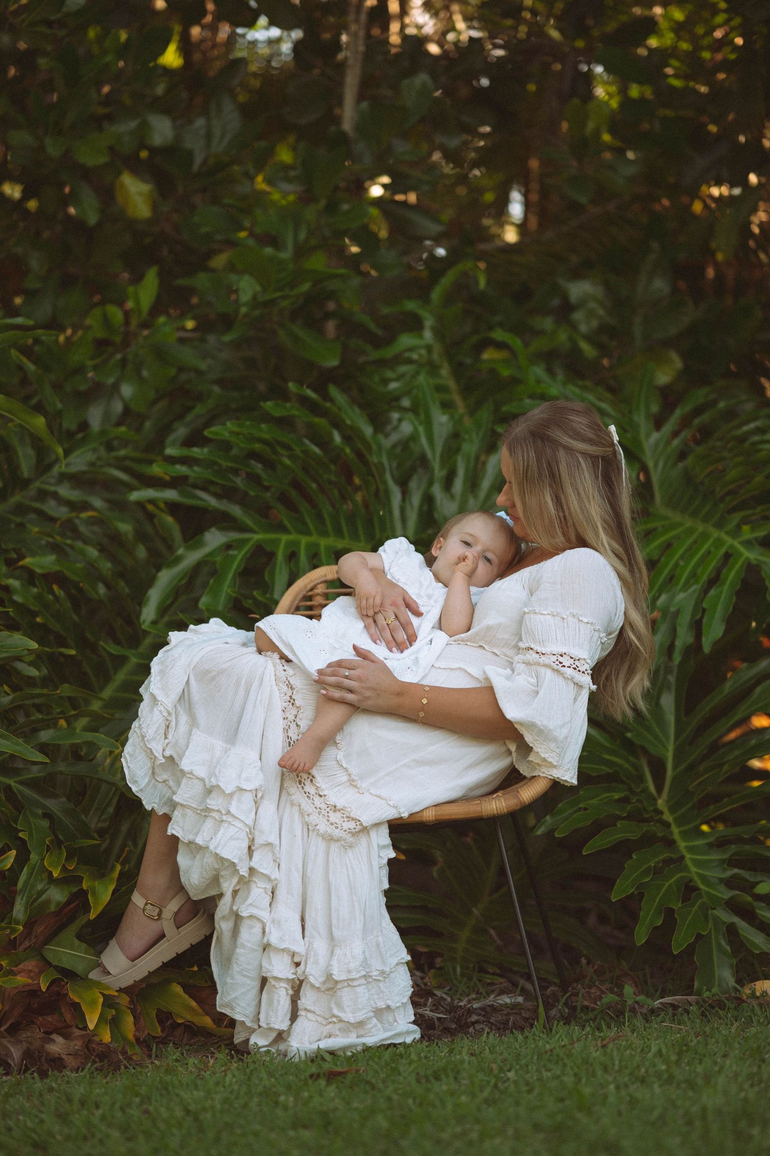 Mother in white dress holding sleeping baby in lush garden setting.