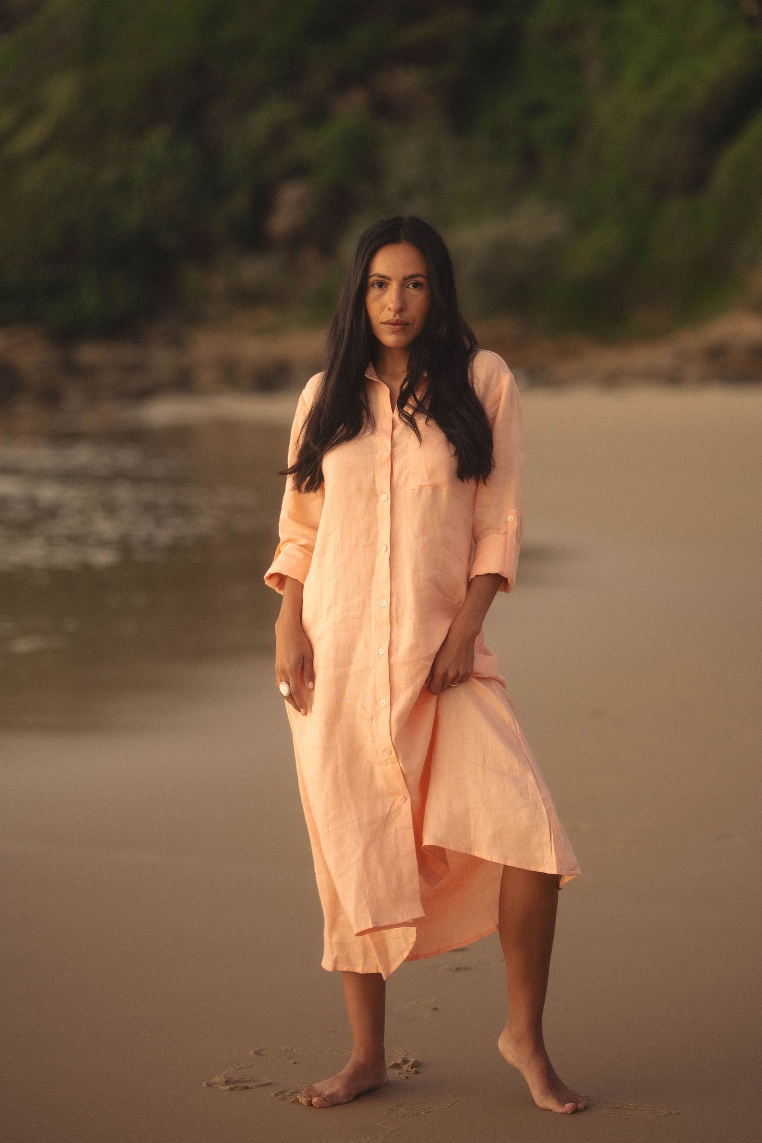 Woman in peach linen shirt dress standing barefoot on sandy beach at golden hour.