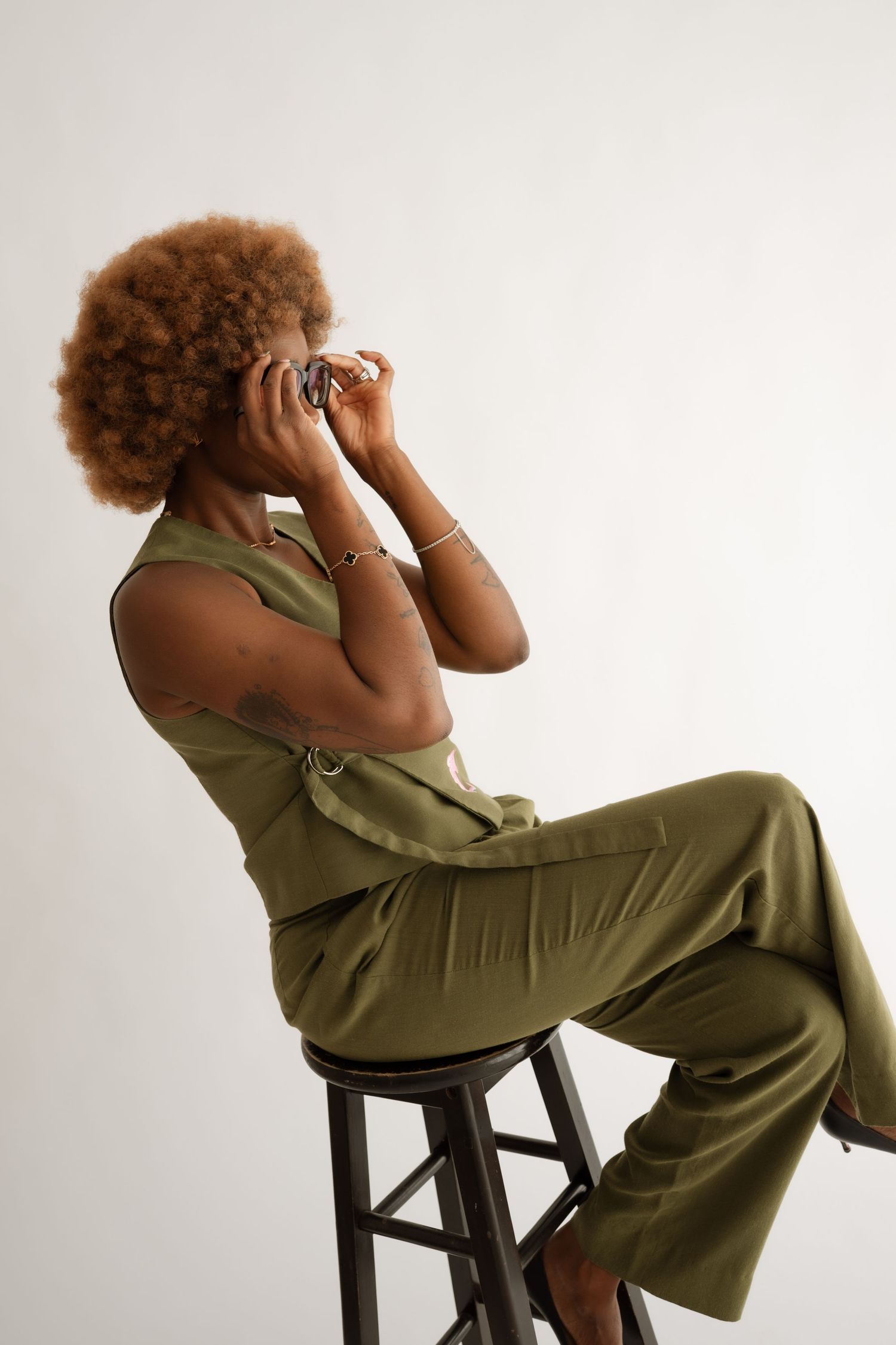 Woman with afro in olive green jumpsuit sitting on stool holding camera to face against white background.