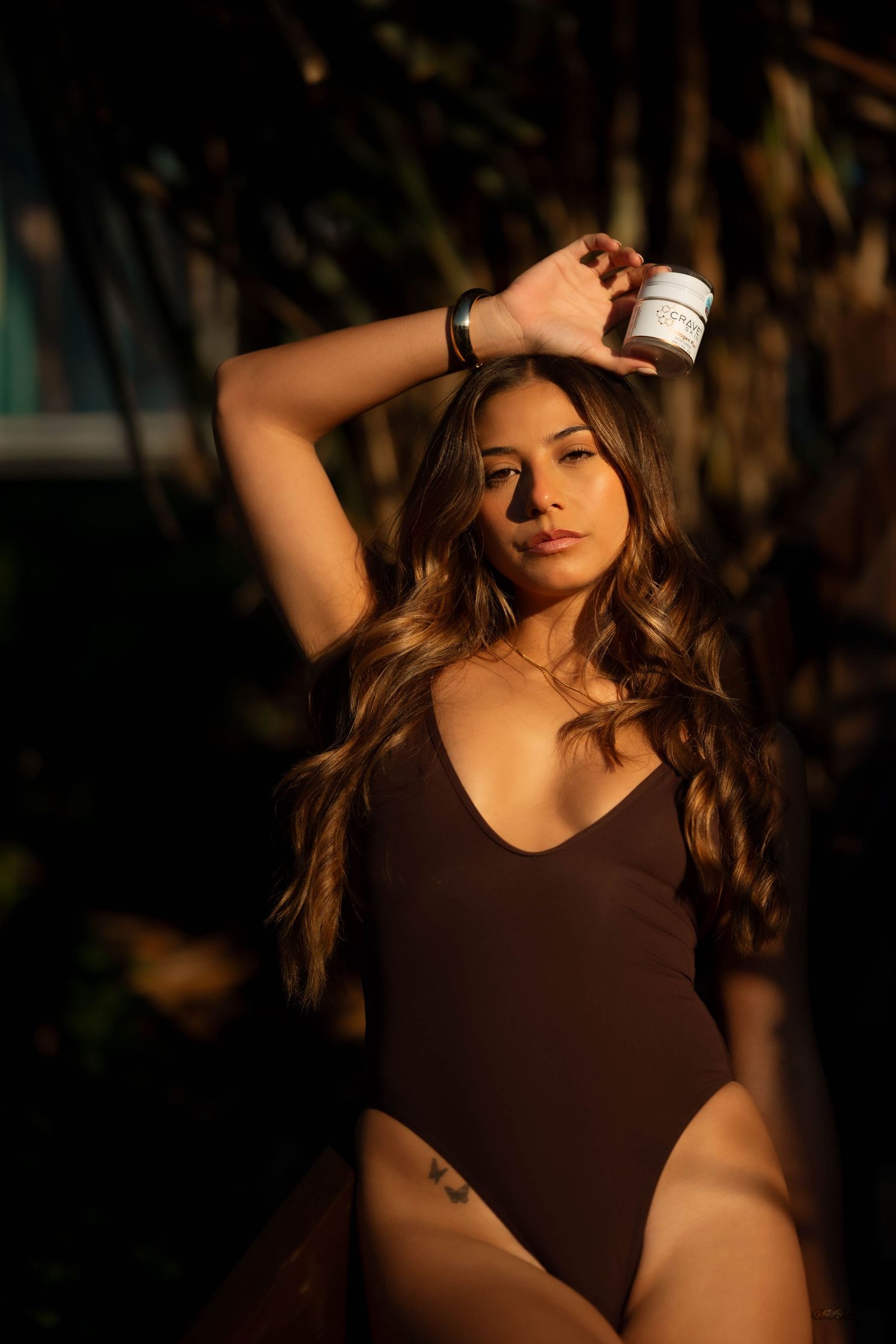 Brunette model in brown bodysuit posing with Crave Skin cream jar outdoors.