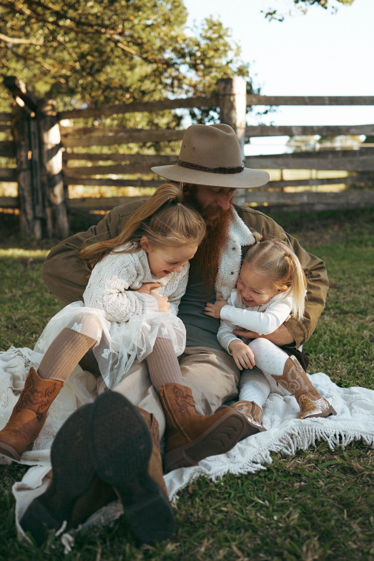Bearded father in hat sitting on blanket with two young daughters at rustic farm fence outdoors.