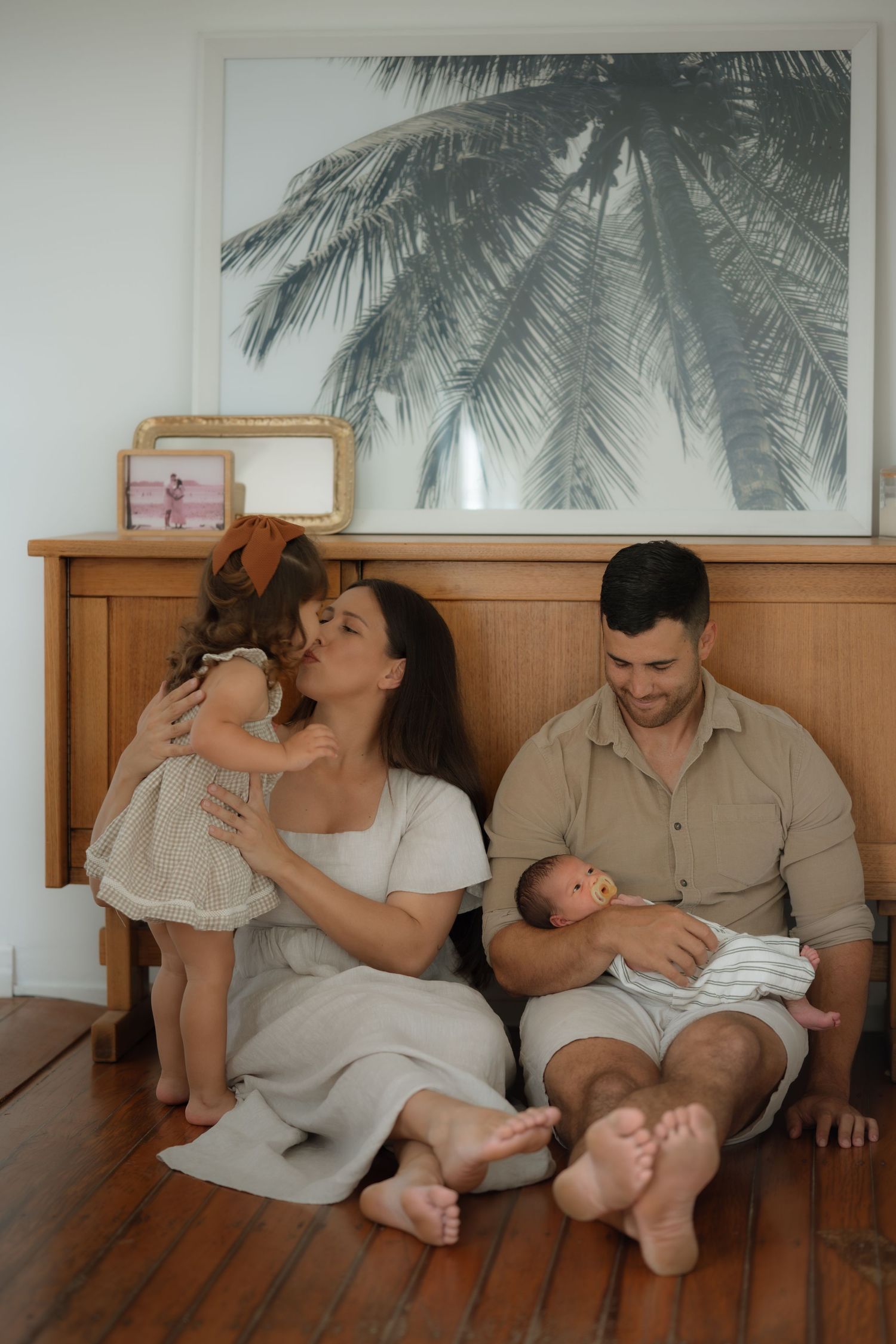 Mother kissing toddler daughter while father holds newborn baby in bright home interior.