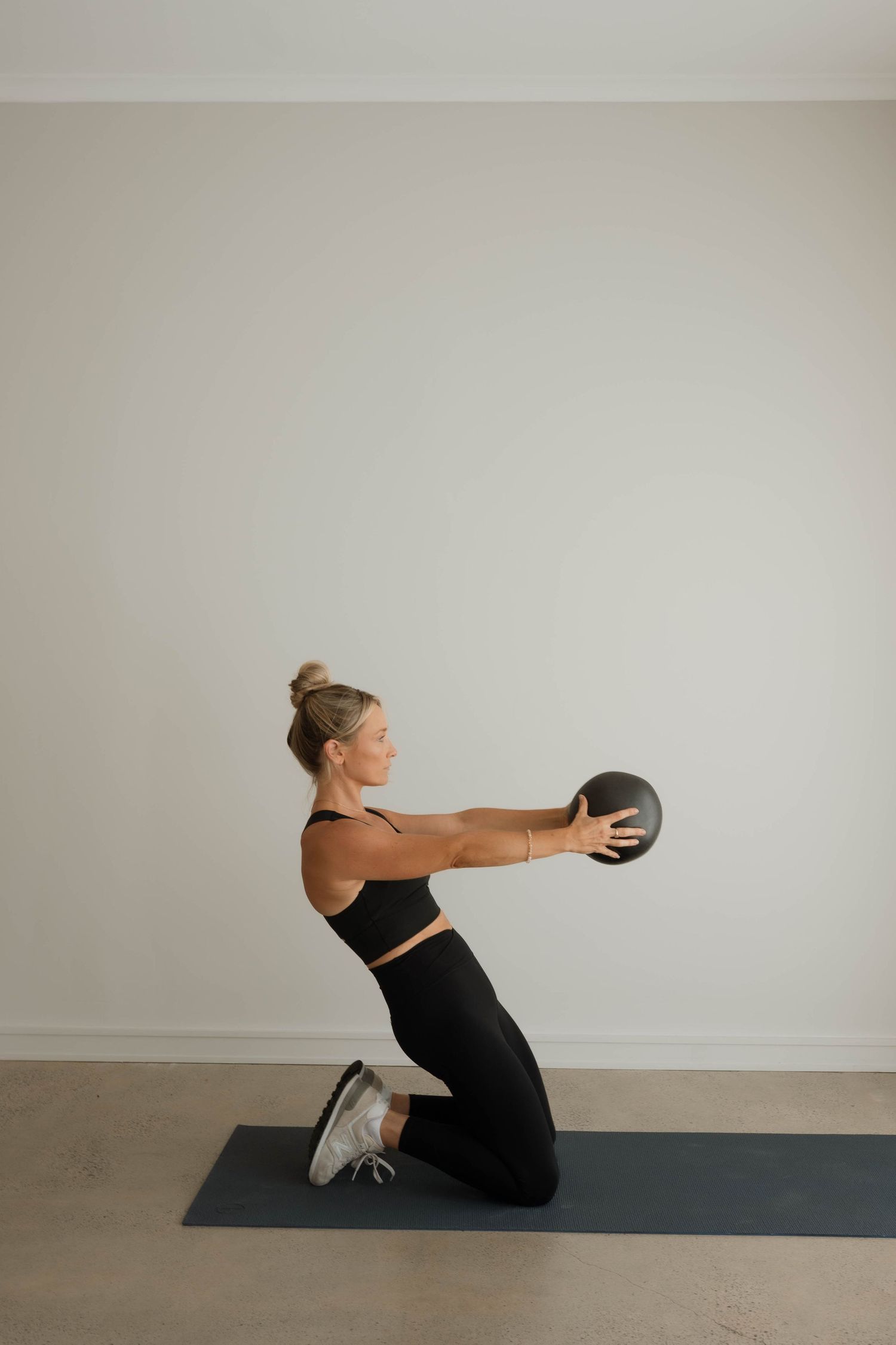 Woman in black activewear kneeling and holding a medicine ball on a yoga mat.