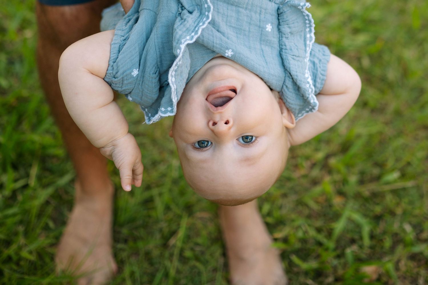 Adorable blue-eyed baby in a teal dress held upside down over green grass, looking up at the camera.