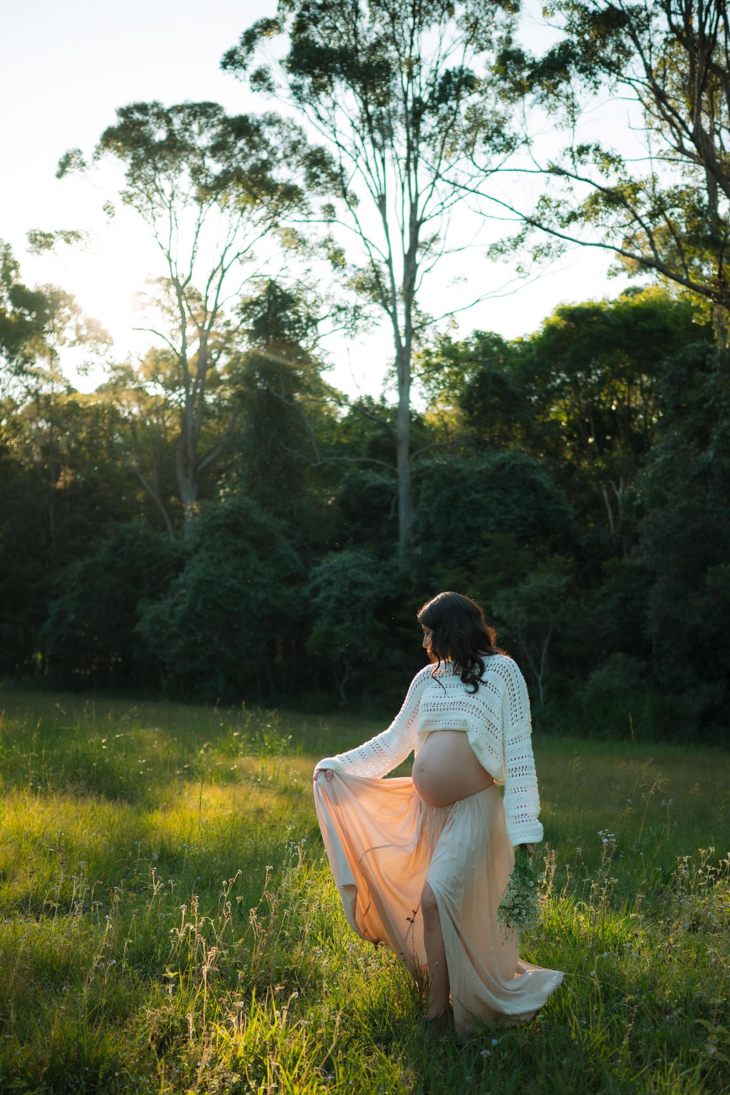 Pregnant woman in white sweater and peach skirt walking away through a lush green field at golden hour.