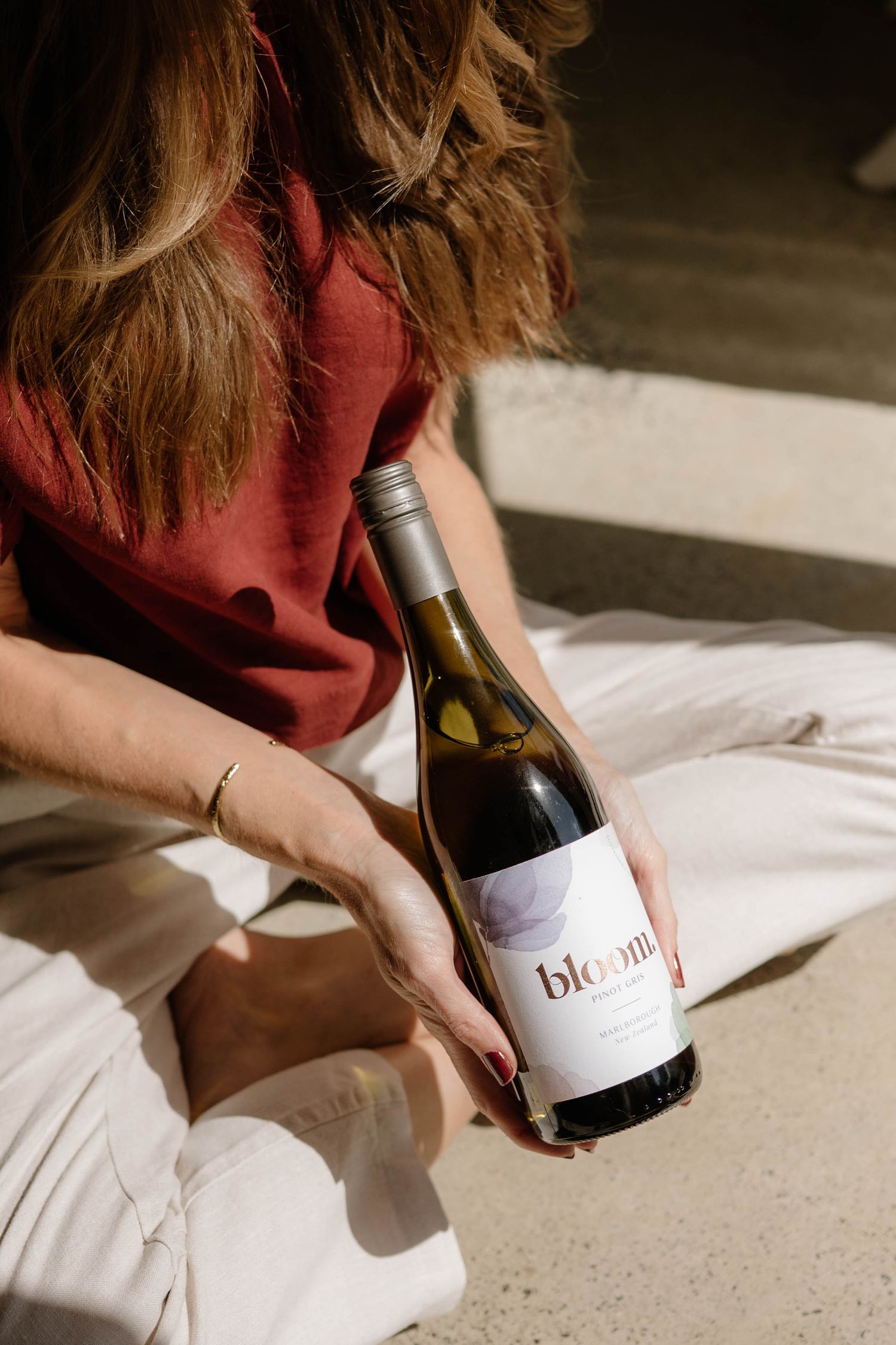 Woman in red top holding Bloom wine bottle in sunlight wearing white pants.