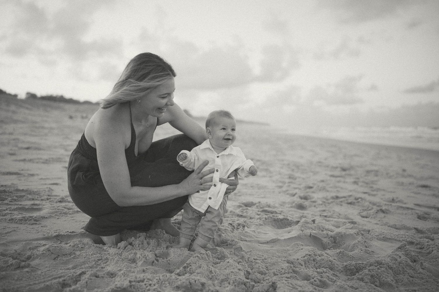 Black and white photo of a mother crouching on sandy beach helping her smiling baby stand in the sand.