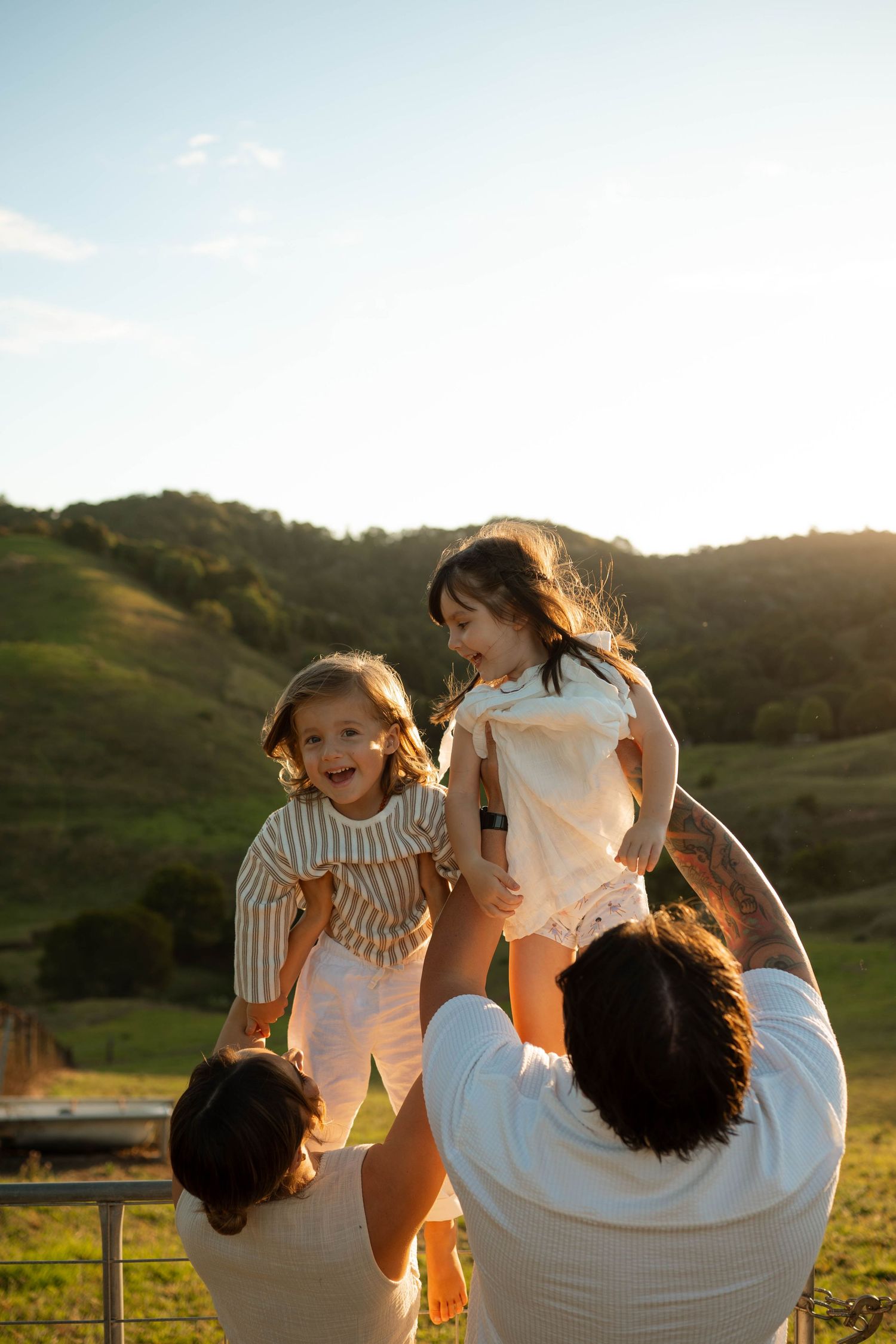 Parents lifting two laughing young daughters in the air against a scenic green hillside at sunset.