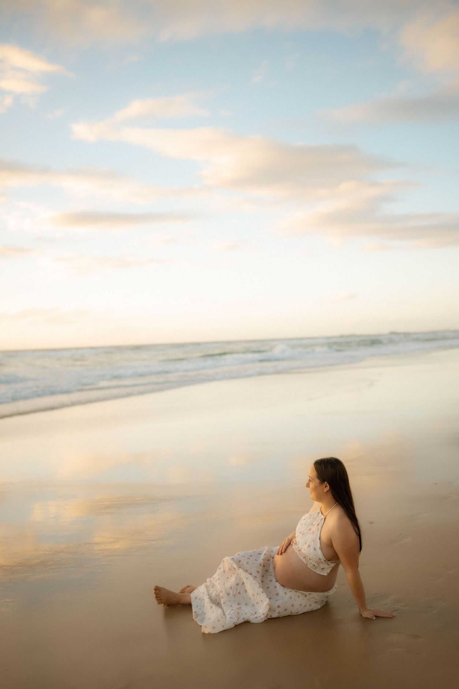 Pregnant woman in white floral dress sitting on wet sand gazing at ocean during golden sunset.