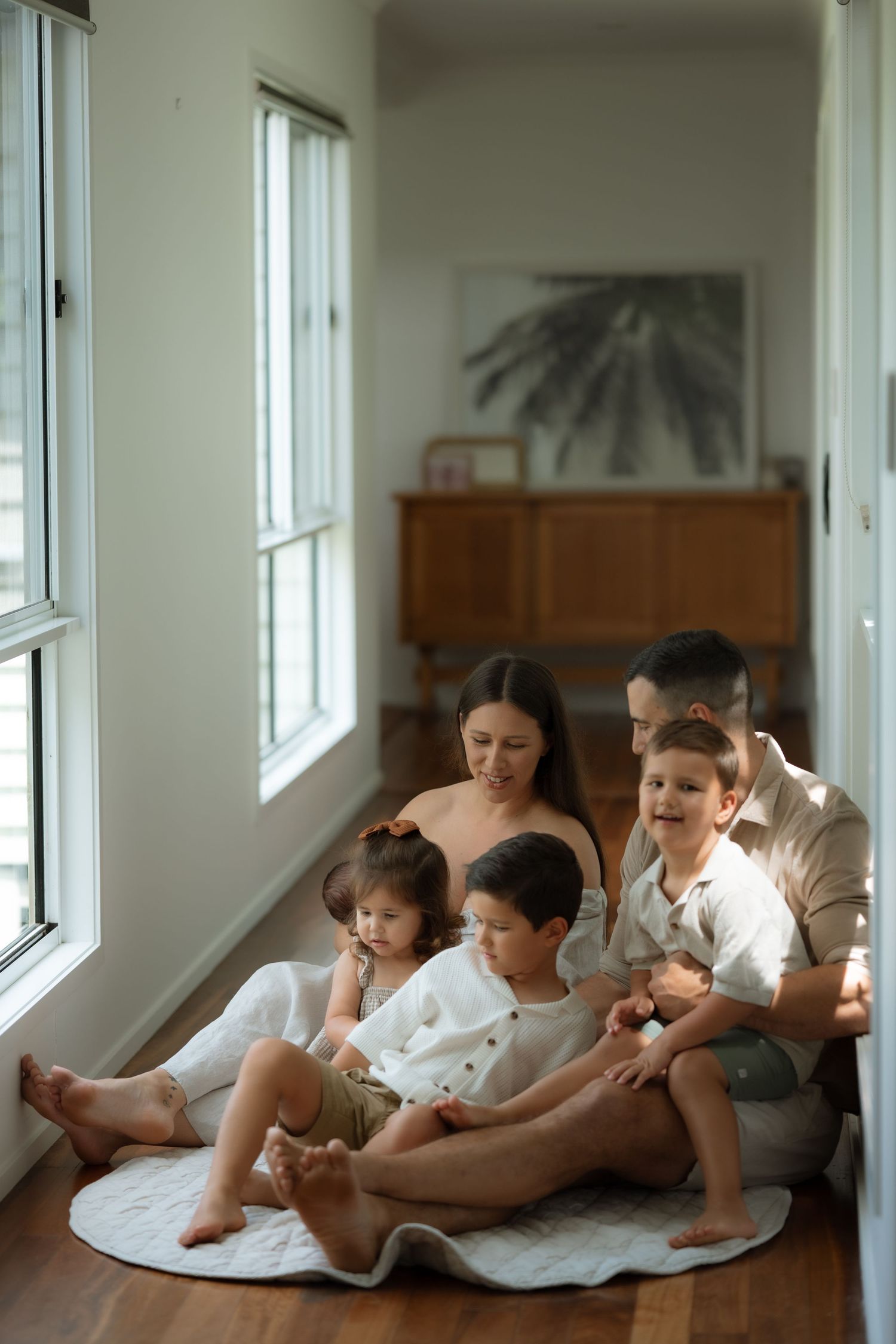 Family of five sitting on floor by bright windows in cozy home, parents with three young children.