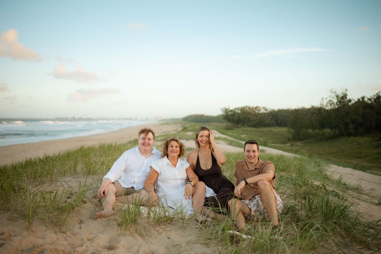 Four adults sitting together on sandy beach dunes with ocean in background.