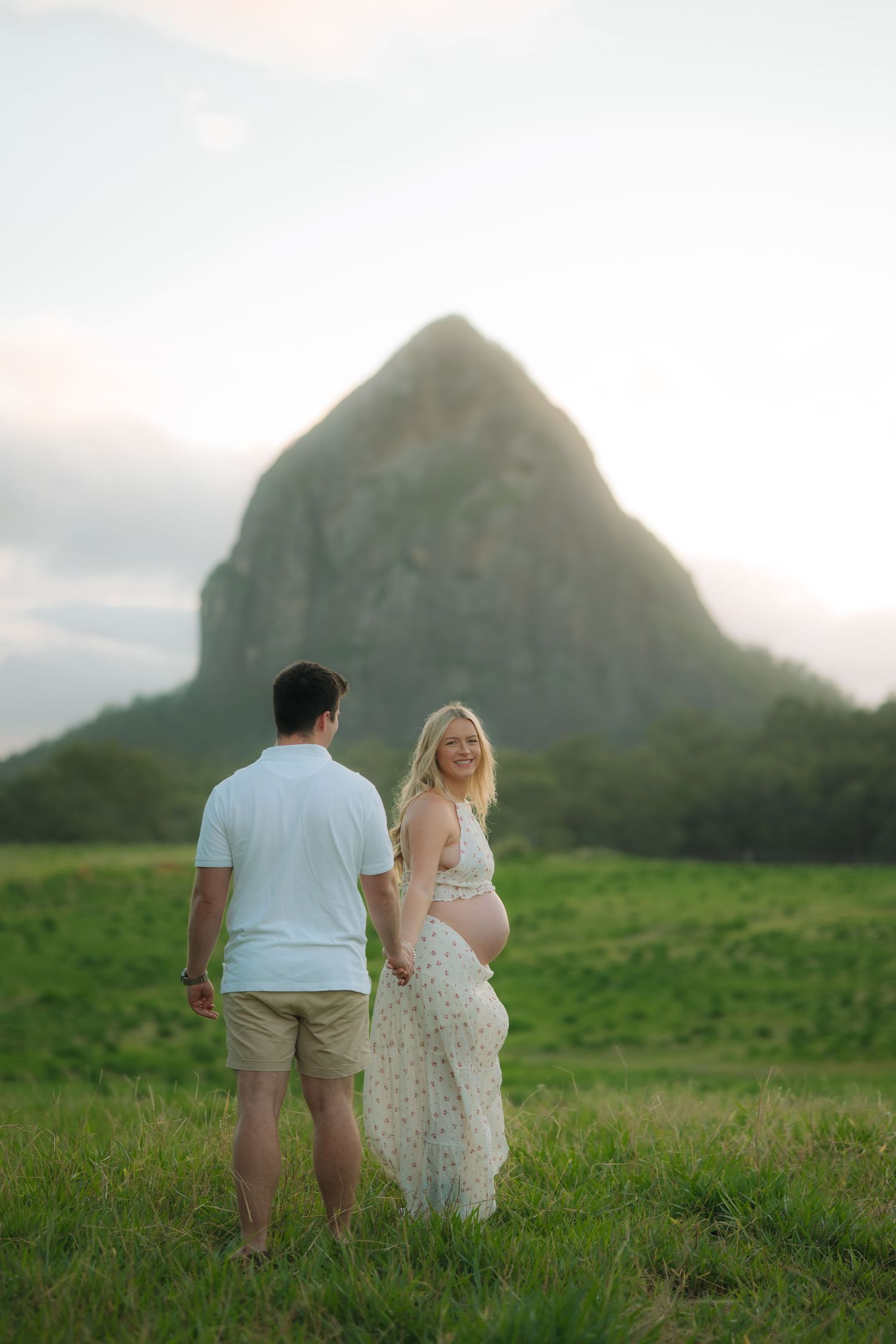 Pregnant woman and partner hold hands in green field, she turns to smile at him with mountain backdrop.