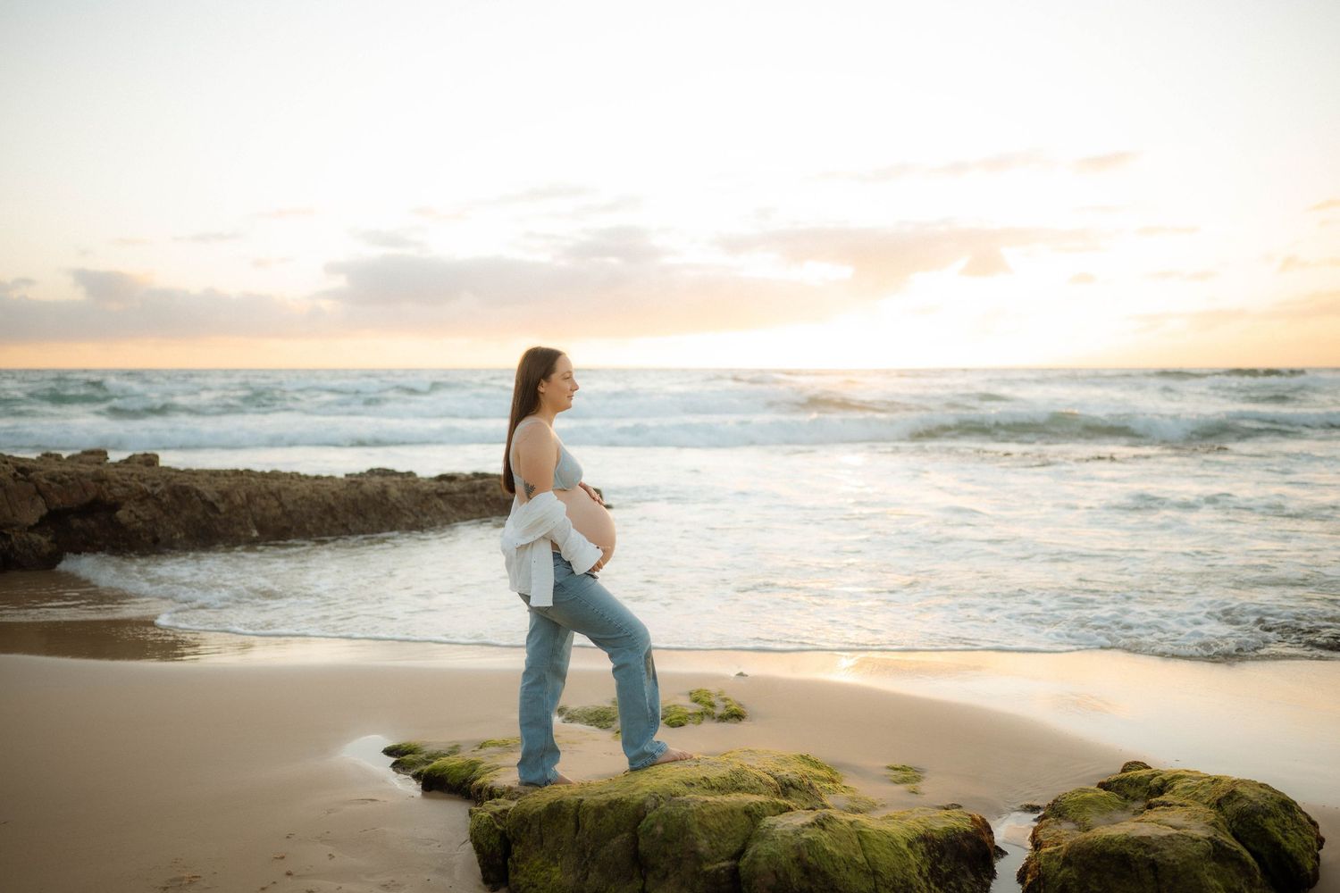 Pregnant woman in jeans and open white shirt standing on mossy rocks at sunset beach.