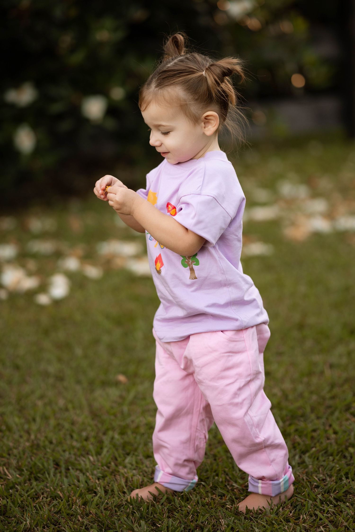 Toddler girl in pink pants and lavender t-shirt playing barefoot on green grass outdoors.