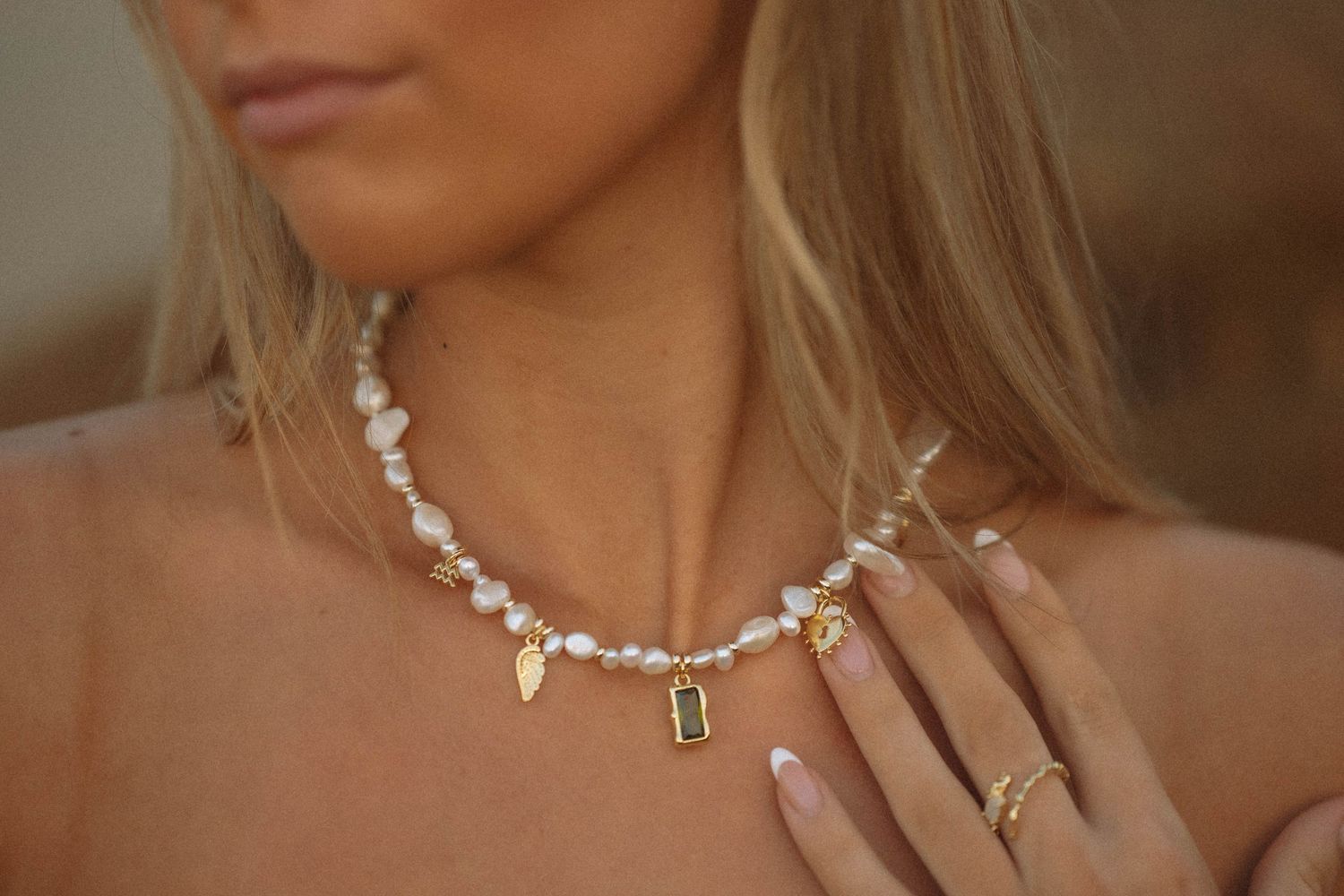 Woman wearing a pearl and gold charm necklace with green gemstone pendant, styled with gold rings and manicured nails.