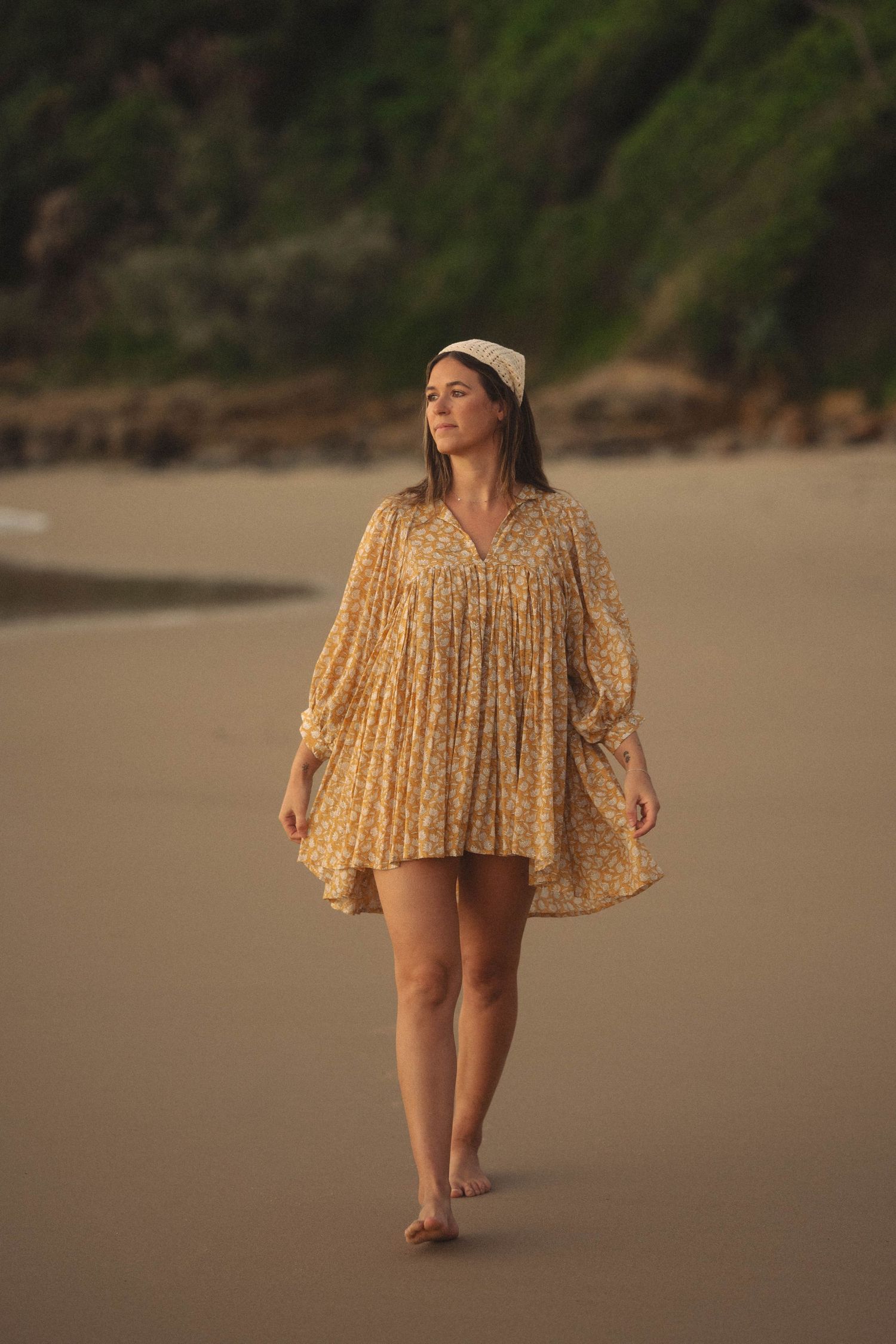 Woman in yellow floral dress walking barefoot on sandy beach at golden hour.